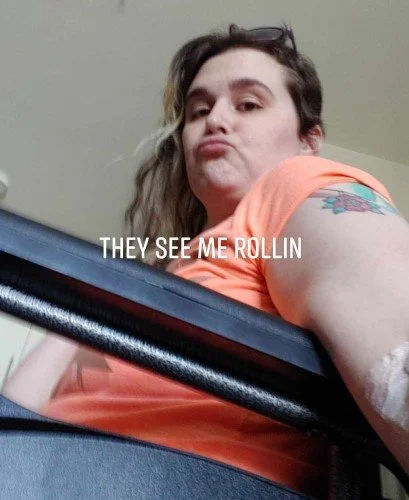 Person with somewhat bleached hair taking a low-angle selfie in a wheelchair with the caption "They See Me Rollin"