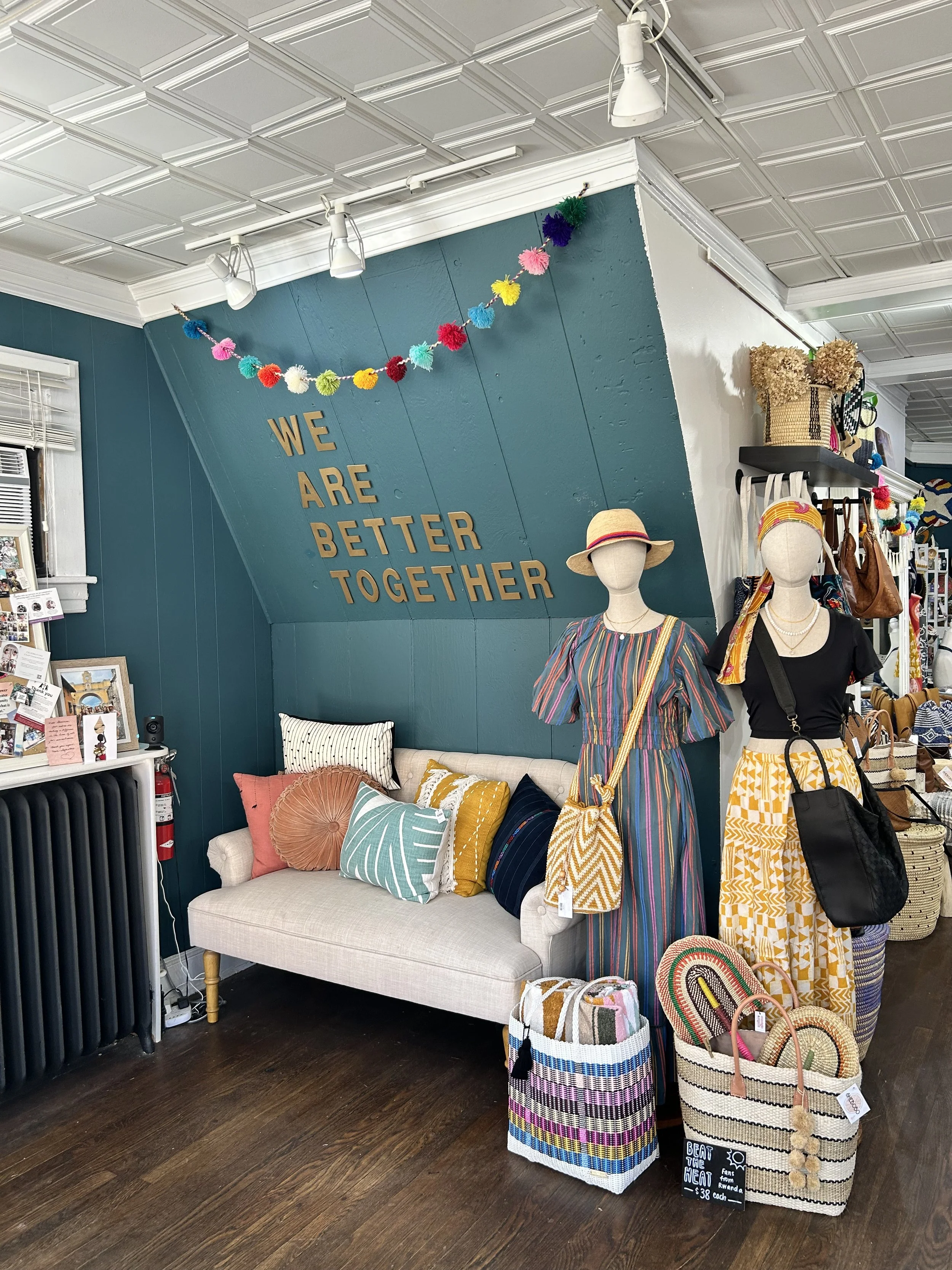 Interior of a boutique store with a teal wall reading "WE ARE BETTER TOGETHER" in gold letters, decorated with a colorful pom-pom garland. Mannequins display summer clothing and hats. A cozy beige sofa with decorative pillows is against the wall, with woven bags placed in front. Various accessories and baskets are on shelves and tables.