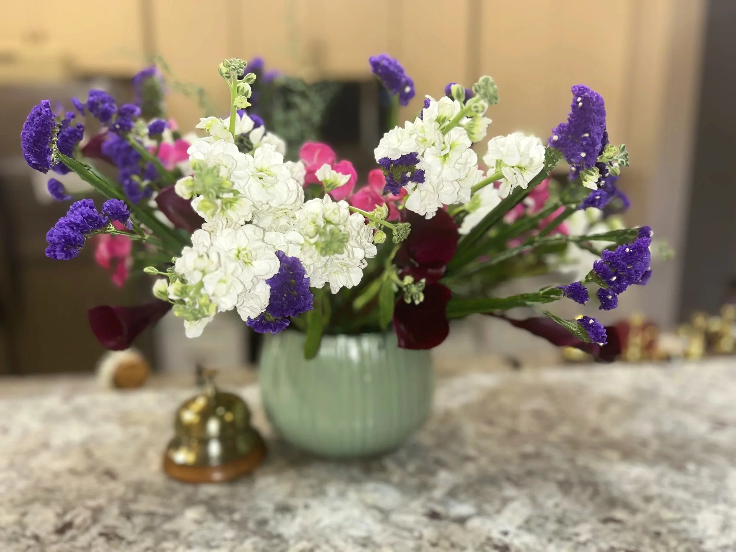 A bouquet of mixed flowers, including white, purple, pink, and dark red blooms in a light green ribbed vase on a granite countertop.