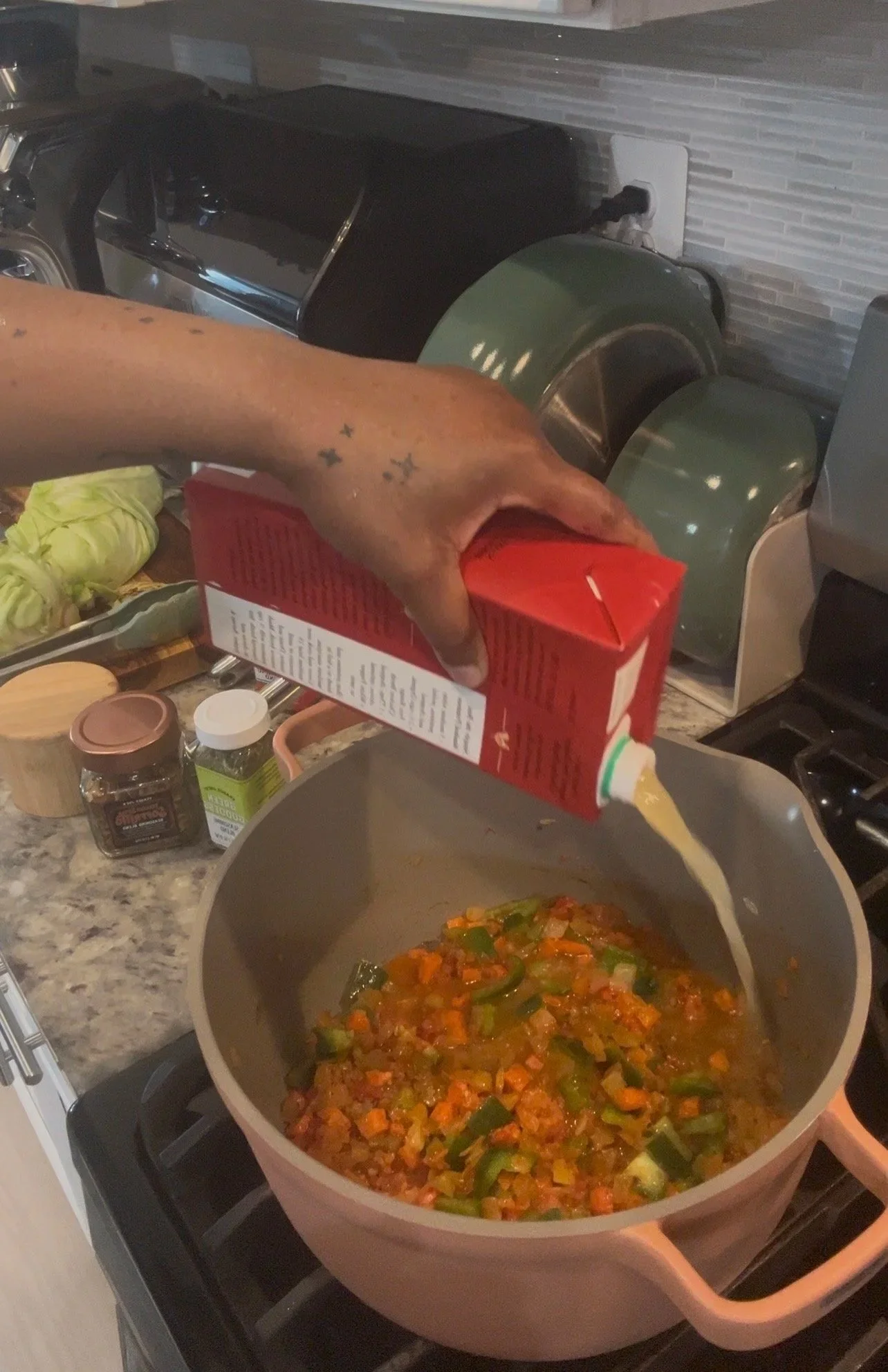 Person pouring creamy soup from a red carton into a large pot on the stove, with chopped vegetables inside the pot.