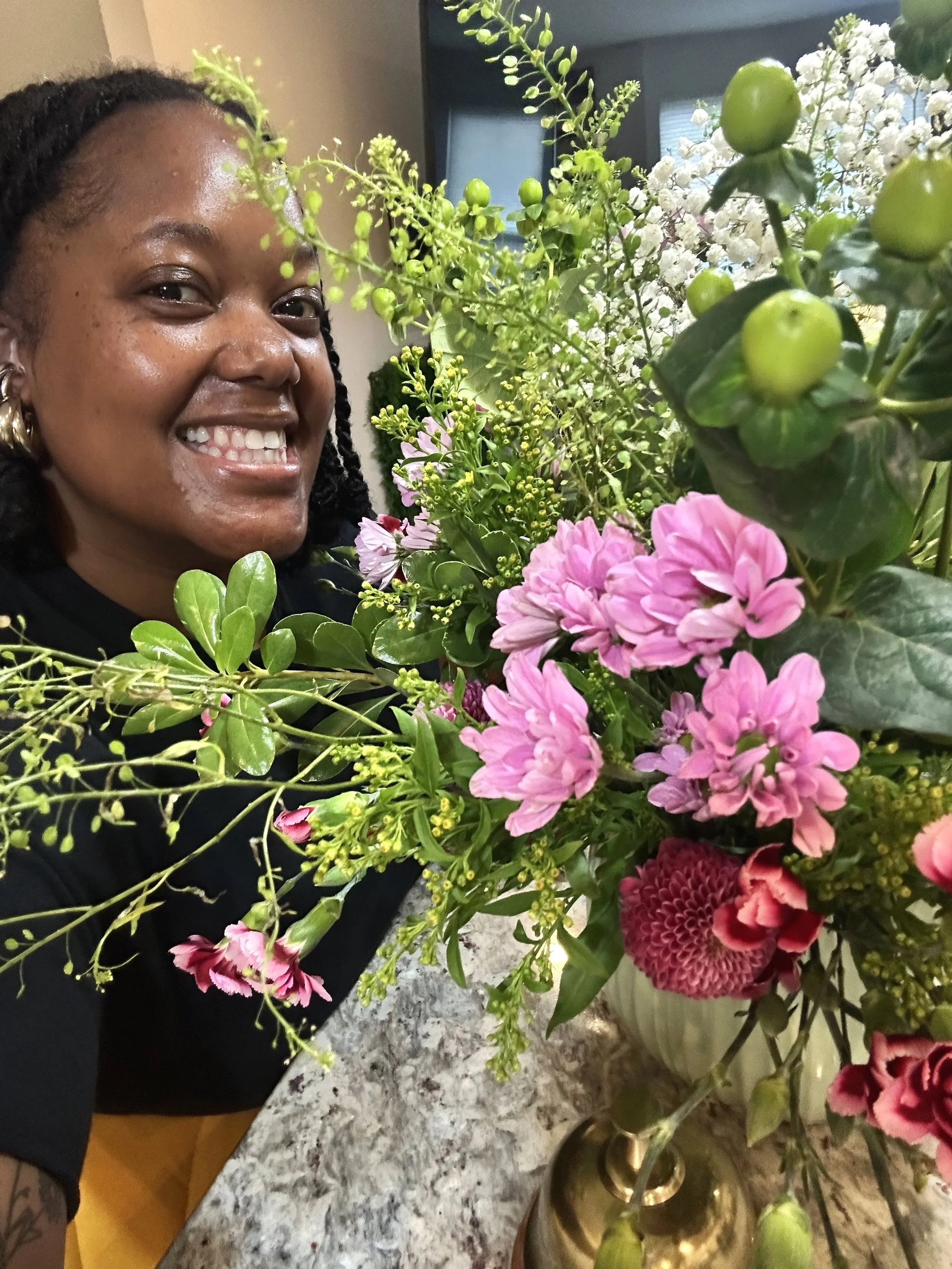 A woman smiling behind a large colorful bouquet of pink, purple, and green flowers in a gold vase on a kitchen counter.
