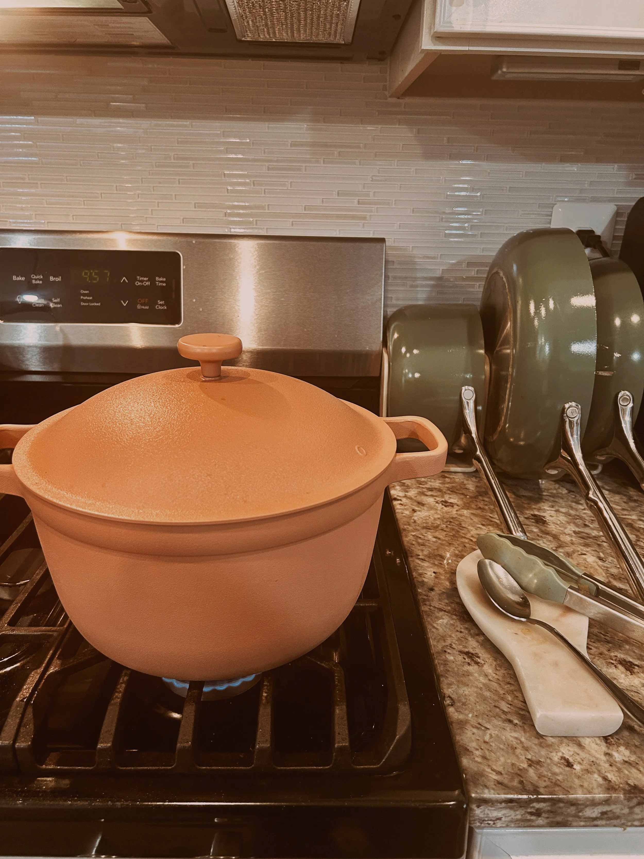 A beige cooking pot on a stove, with a lid on and steam coming out, in a kitchen. To the right, there are kitchen utensils including tongs and a spoon on a plate, on a granite countertop. In the background, there is a textured tile backsplash and a microwave oven.