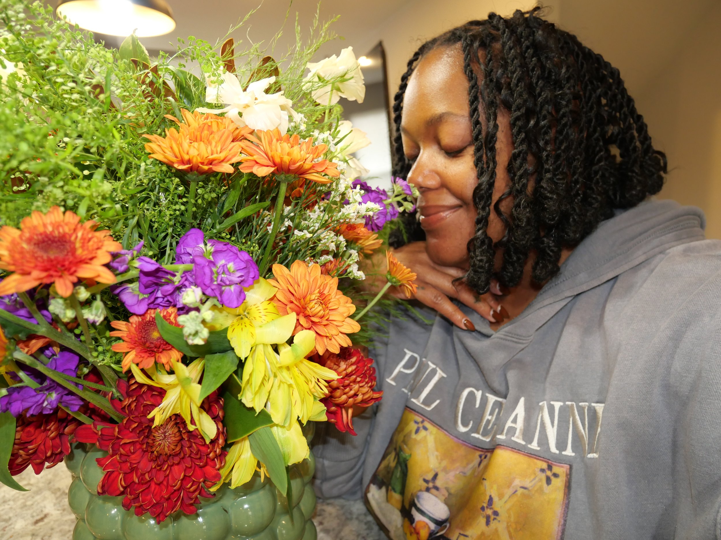 A woman with dark, curly hair smelling a vibrant bouquet of mixed flowers in a green vase.