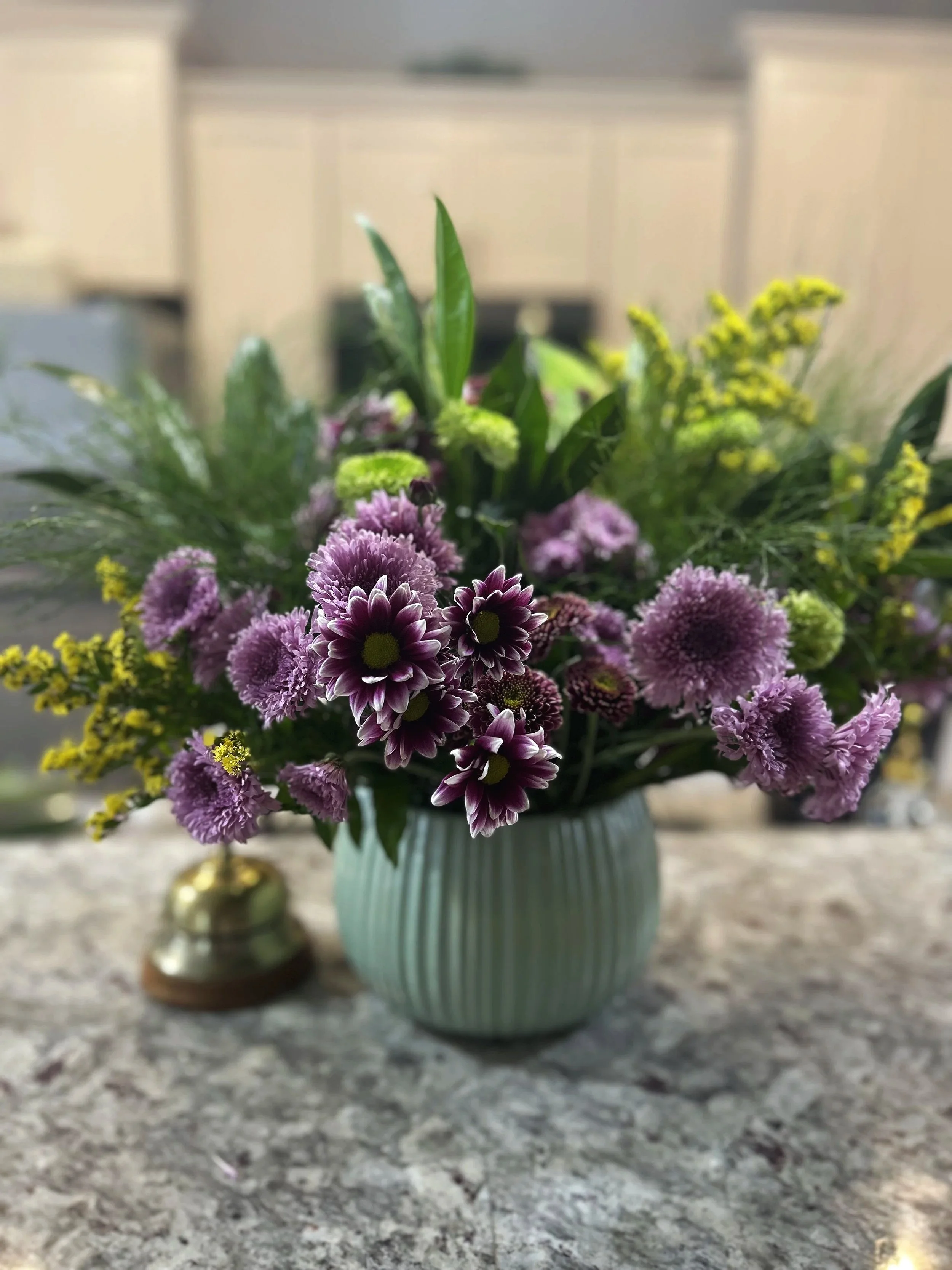 A vase of purple and green flowers on a gray marble countertop with blurred background.