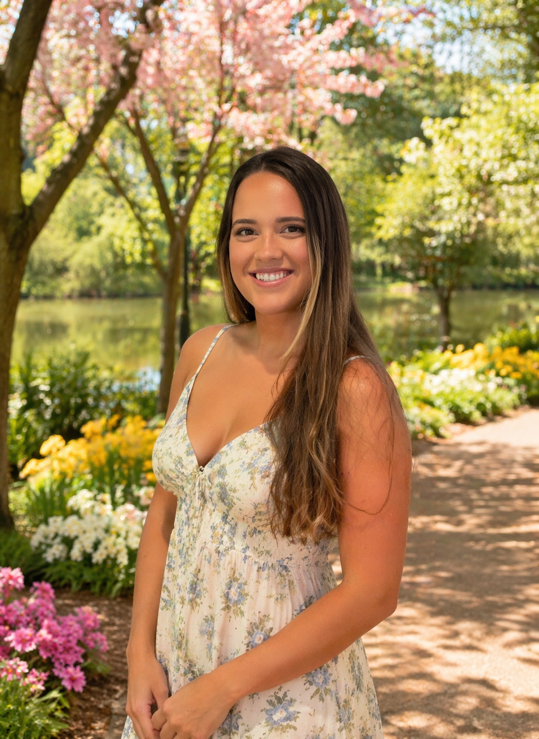 A young woman with long brown hair wearing a light-colored, floral summer dress standing on a garden path with colorful flowers and trees behind her, by a pond, on a sunny day.
