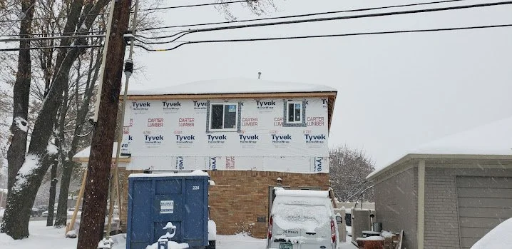 Two-story house under construction with Tyvek house wrap, two small windows on the second floor, a brick foundation, a blue dumpster, and a white vehicle covered in snow in a snowy neighborhood.