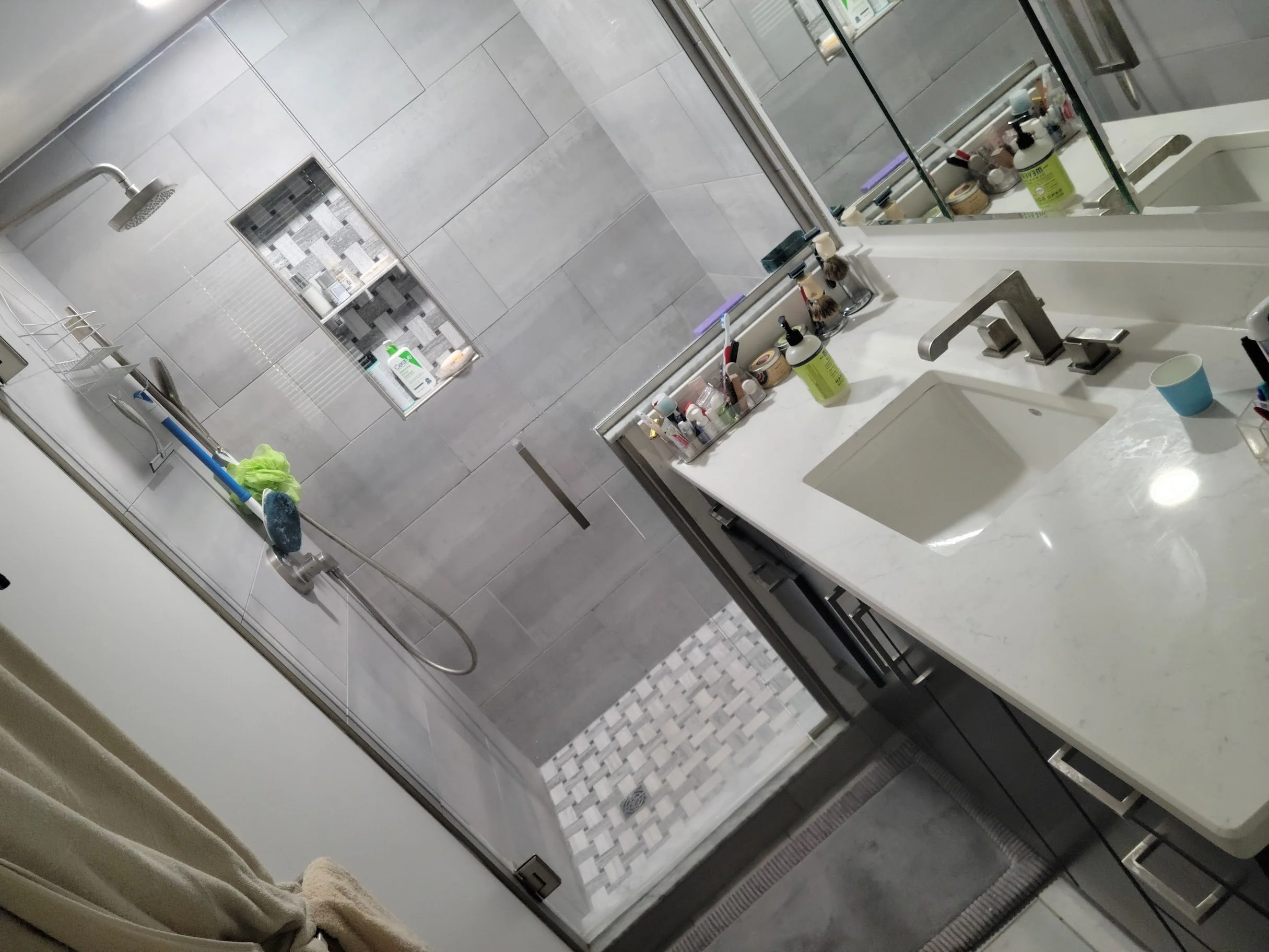 Modern bathroom with gray tiled walls and a glass shower door. Inside the shower, there is a showerhead, a small built-in shelf with toiletries, and a cleaning mop with a green cloth hanging over it. Outside the shower, there is a white bathroom vani