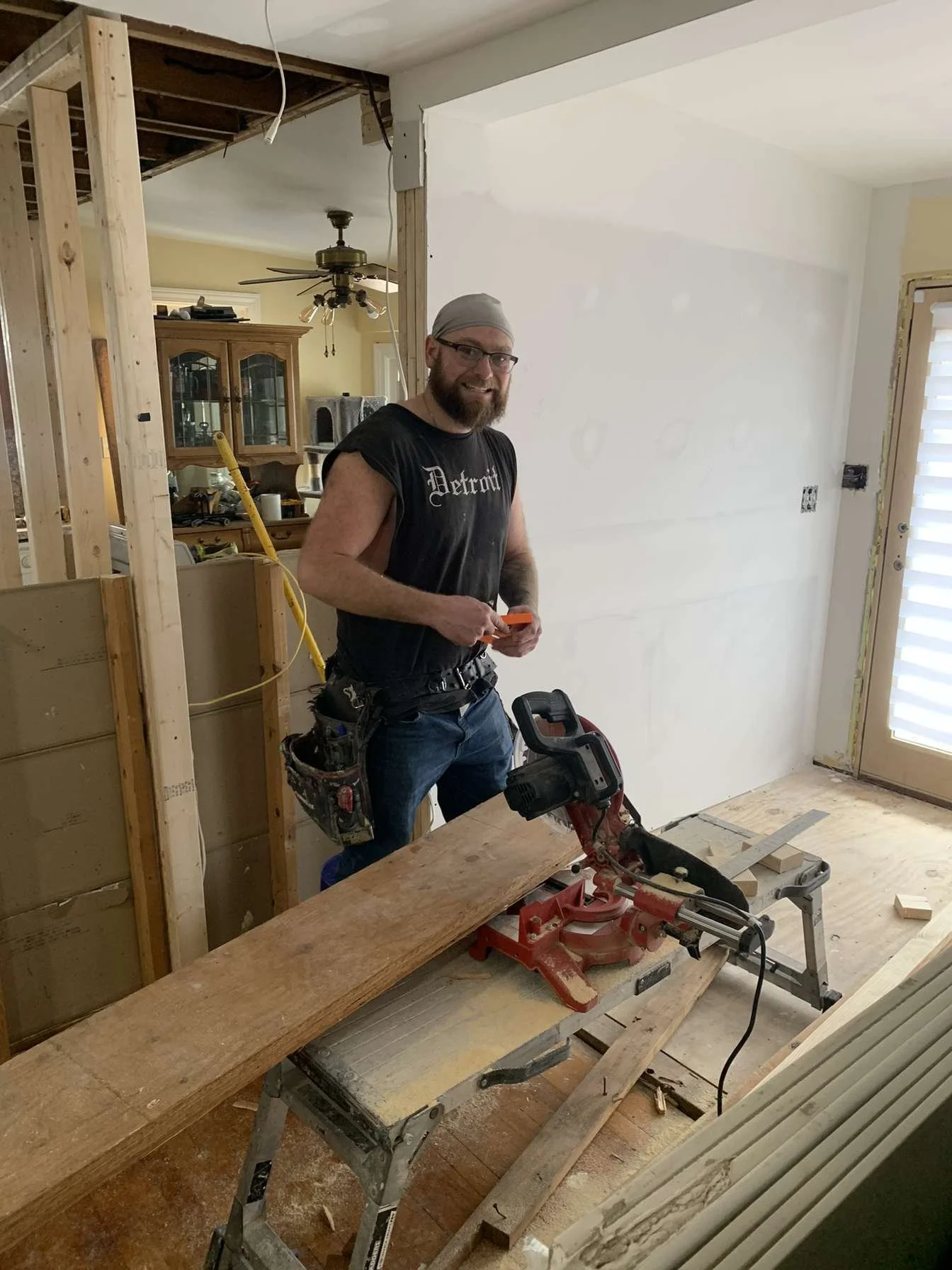 A man with a beard and glasses, wearing a gray bandana and a black T-shirt, working inside a house under renovation. He stands beside a red miter saw on a wooden workbench, holding an orange object and smiling at the camera. The room has unfinished d