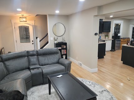 Living room with gray sectional sofa, black coffee table, and a round mirror on the wall, connected to a kitchen with black cabinets and wooden countertops.