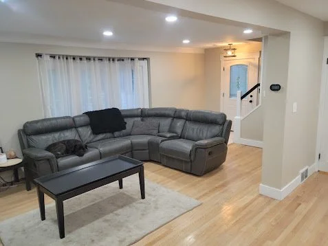 Living room with a large gray sectional sofa, black throw pillows, a black coffee table, a beige area rug, and a window with white curtains.