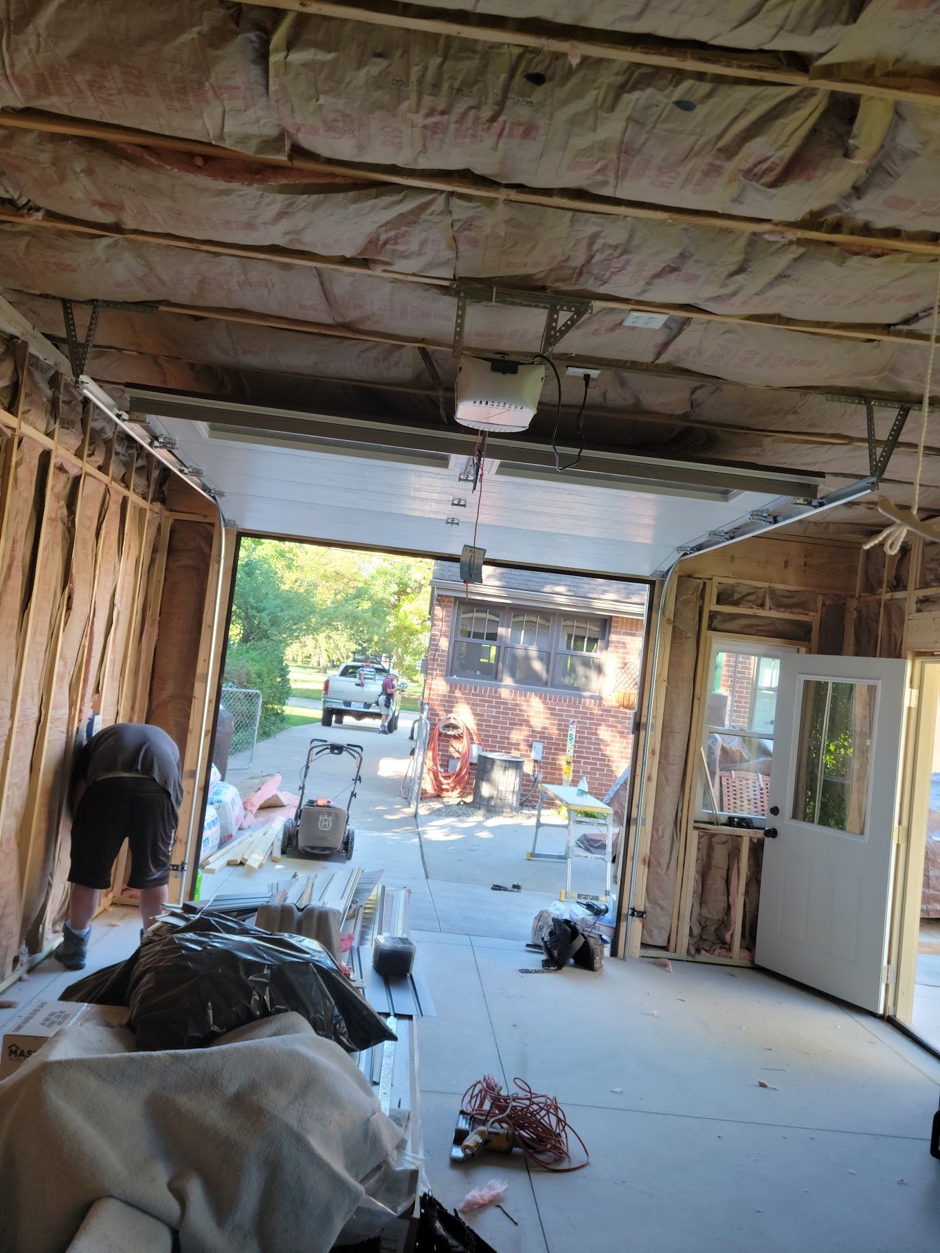 Construction inside a garage with the garage door open, showing a driveway, a person working near the wall, and various tools and materials scattered on the floor.