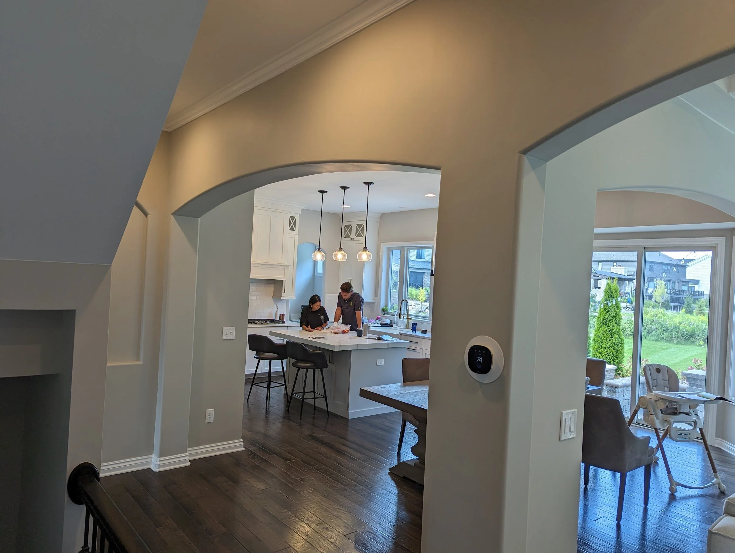 Interior view of a modern kitchen and dining area in a home with large windows showing an outdoor scene with greenery, white cabinetry, pendant lights, and people working at the kitchen island.