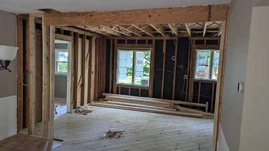 A room under construction with exposed wall studs and a wooden ceiling beam, three windows, and wooden floorboards.