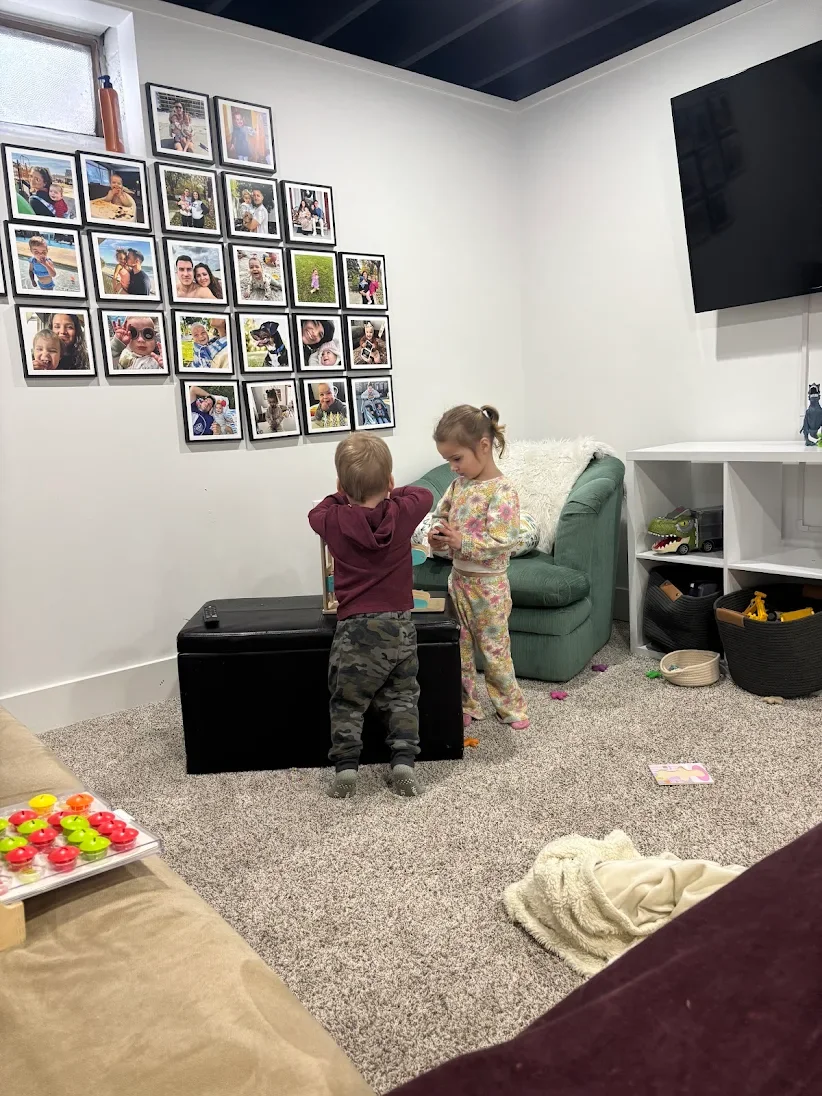 Two young children, a boy and a girl, are standing near a green sofa in a living room, engaged in an activity. The wall behind them displays a collage of family photos in black frames. To the right, there is a white shelving unit with toys, and a large flat-screen TV is mounted on the wall. The carpeted floor has various scattered toys and items.