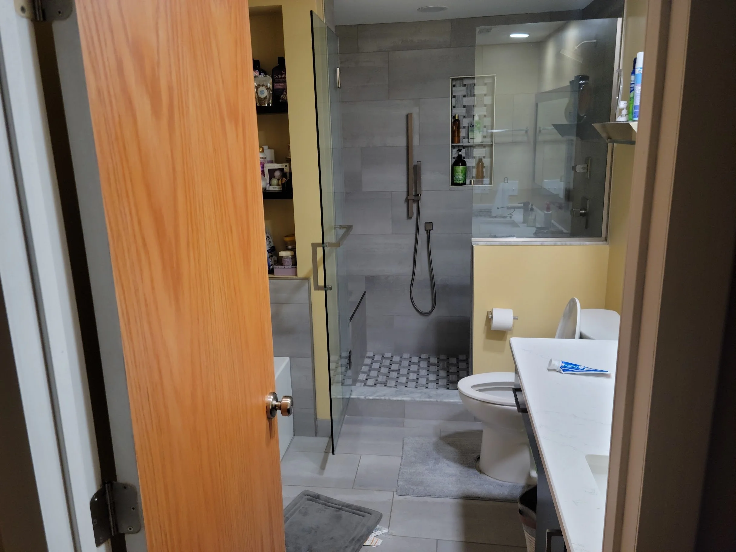 Bathroom with a glass shower enclosure, gray tiled walls, a small shelf with toiletries, a white toilet, and a white countertop with a blue and white toothpaste tube.