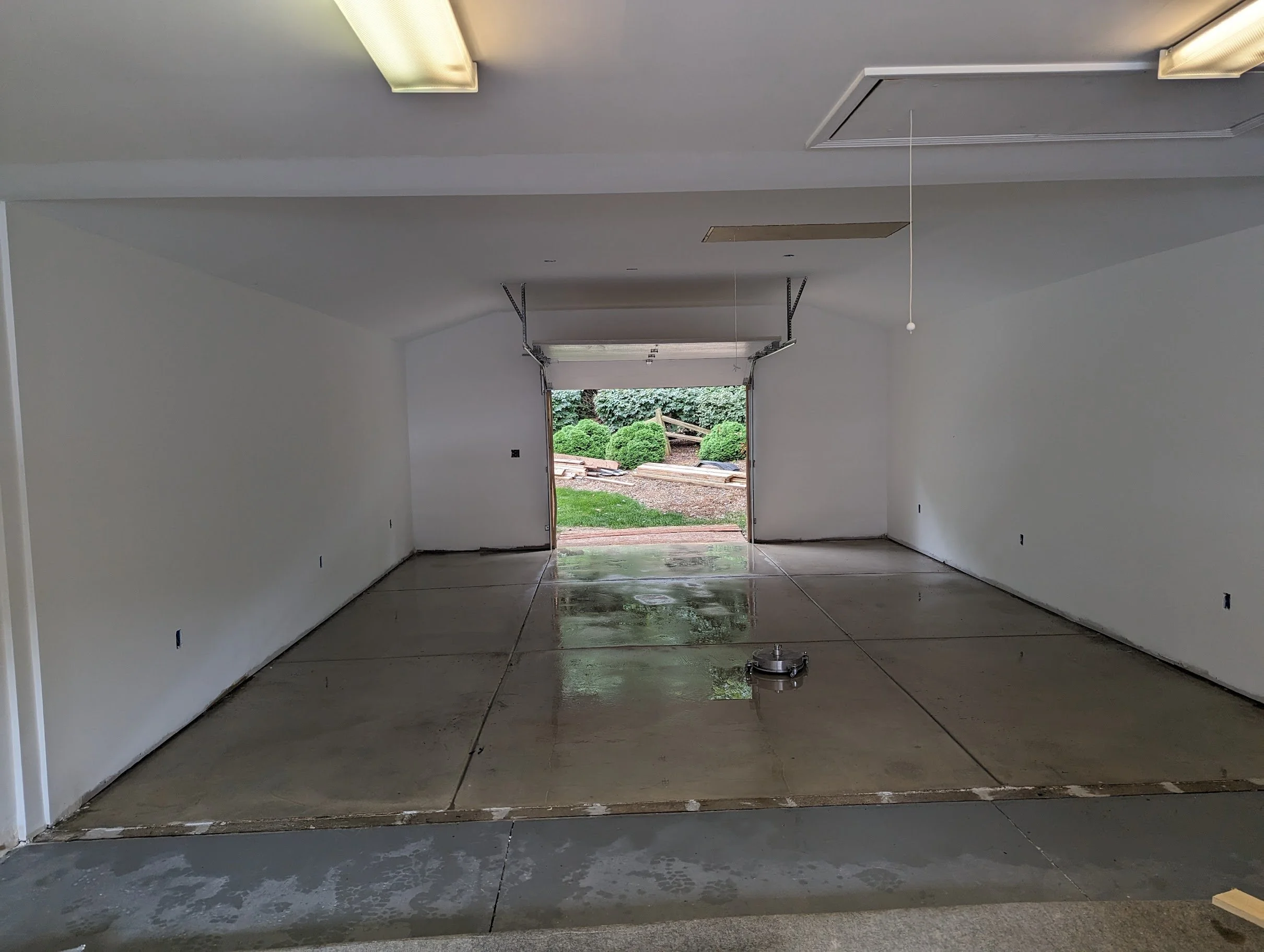 Empty garage with a view of the backyard, showing a green lawn, bushes, and construction materials outside.