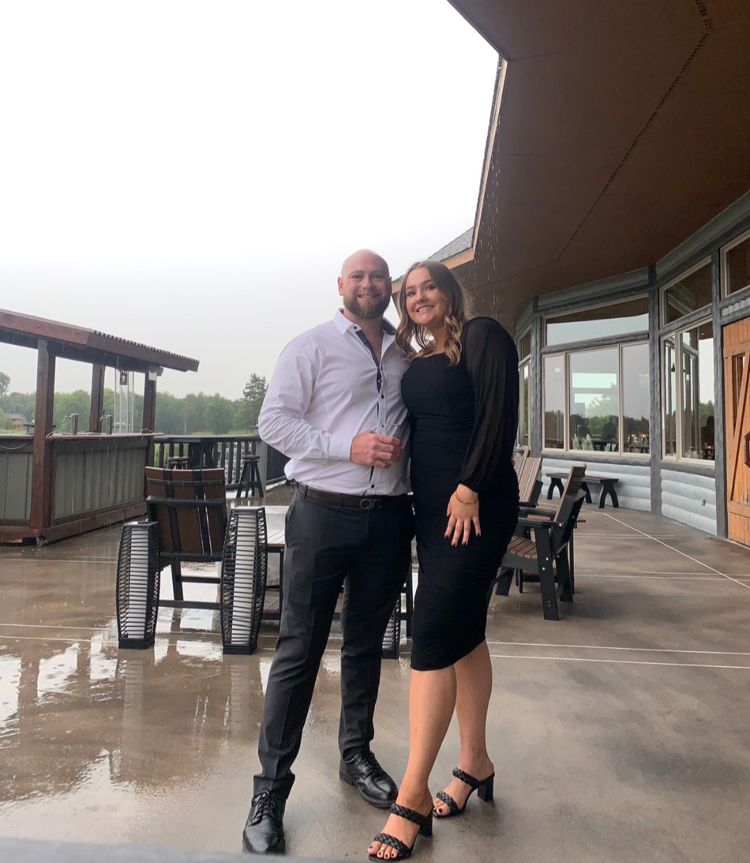 A man and woman standing close together on a wet outdoor patio, smiling. The man is wearing a white shirt and dark pants, holding a glass, and the woman is wearing a black dress and heels. They are near a building with large windows and outdoor tables, with a cloudy sky overhead.