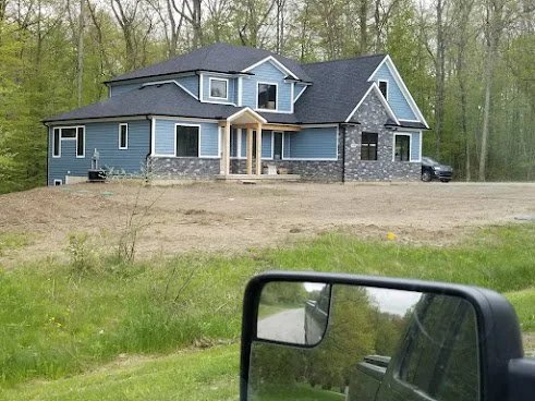 A large blue house with gray stone accents, surrounded by trees, viewed from the side of a road with a car's side mirror in the foreground.