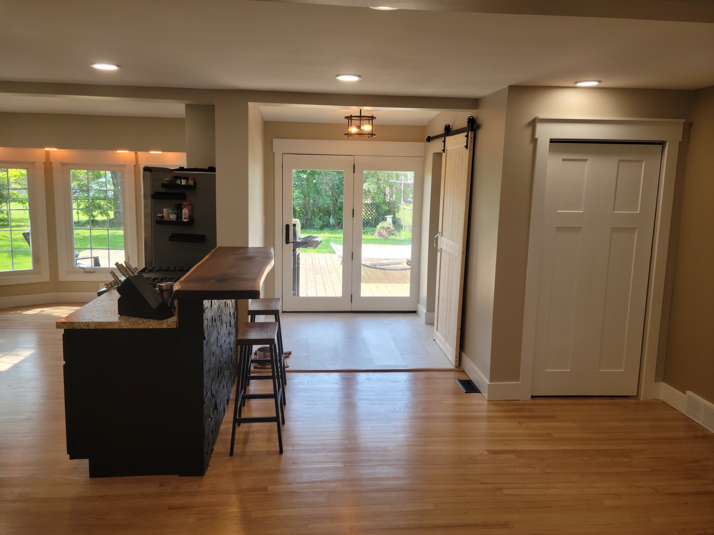 A view of a living room with a kitchen island, bar stools, and several windows showing a green backyard.