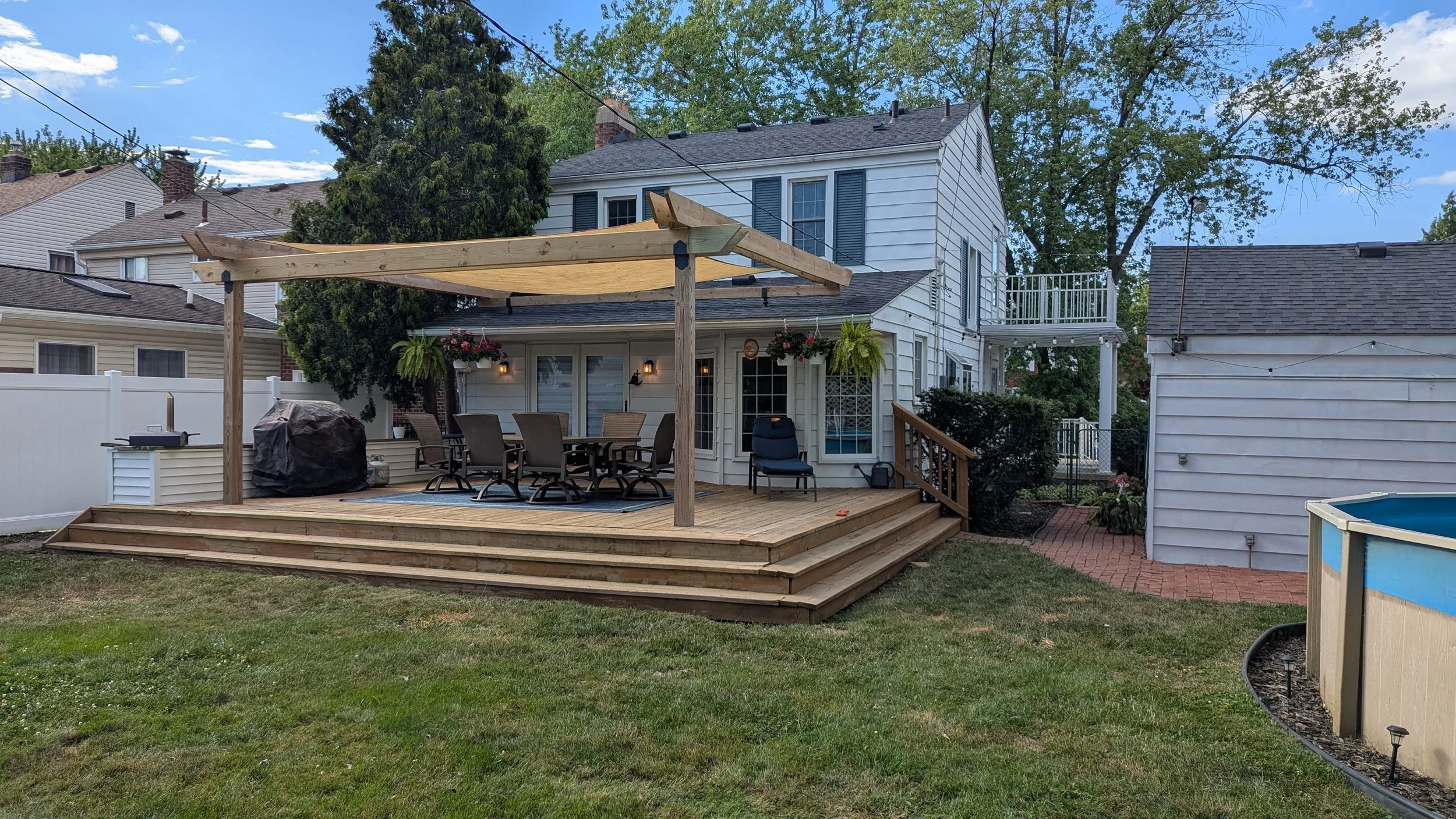 A backyard patio area with a wooden deck, outdoor dining table with six chairs, a covered area with a canopy, hanging flower baskets, a grill, and a backyard fence. There are neighboring houses and trees in the background.