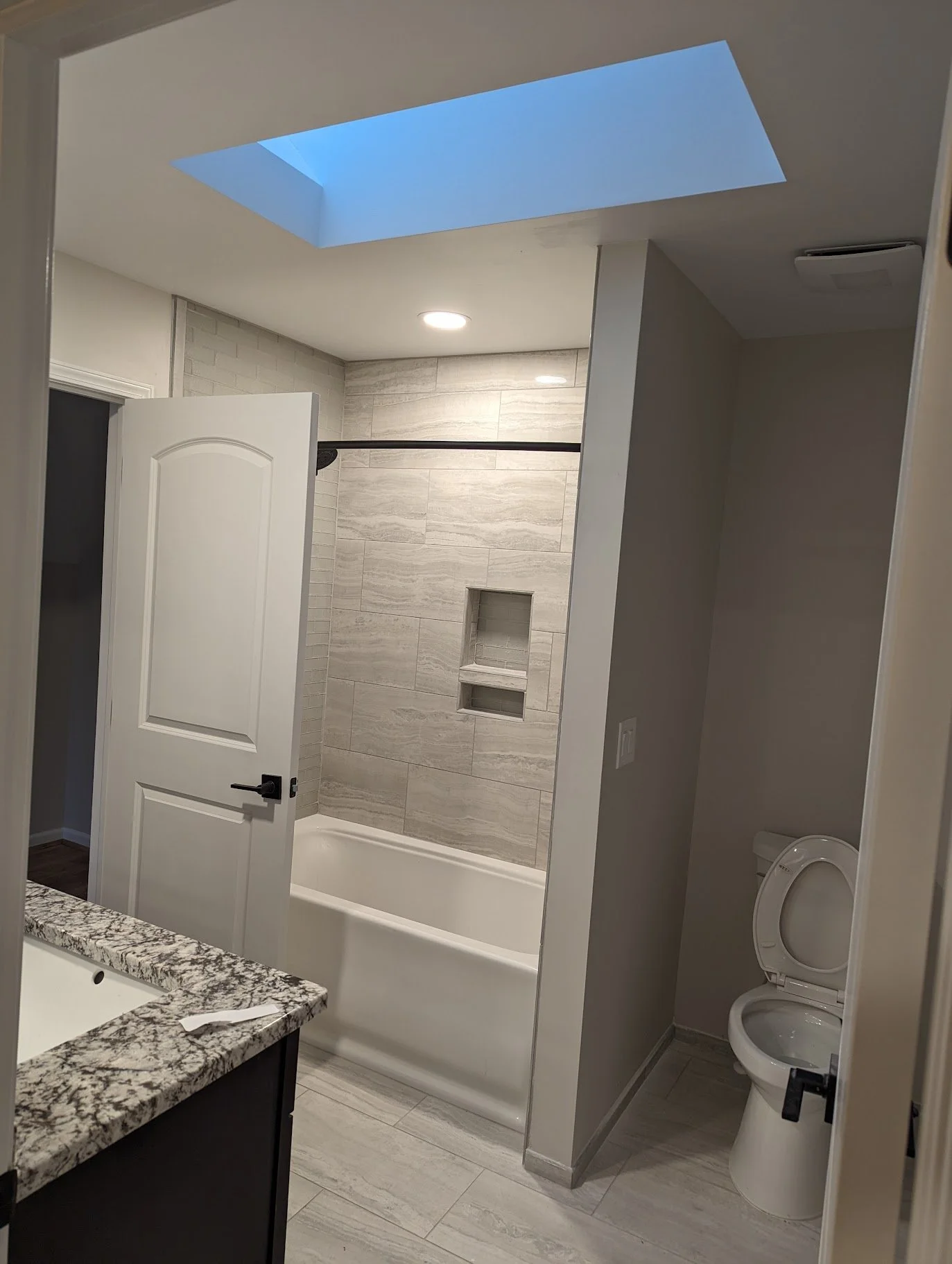 Bathroom with a bathtub and shower area, a wall with beige tiles, a skylight in the ceiling, a toilet, and a granite countertop with a sink.