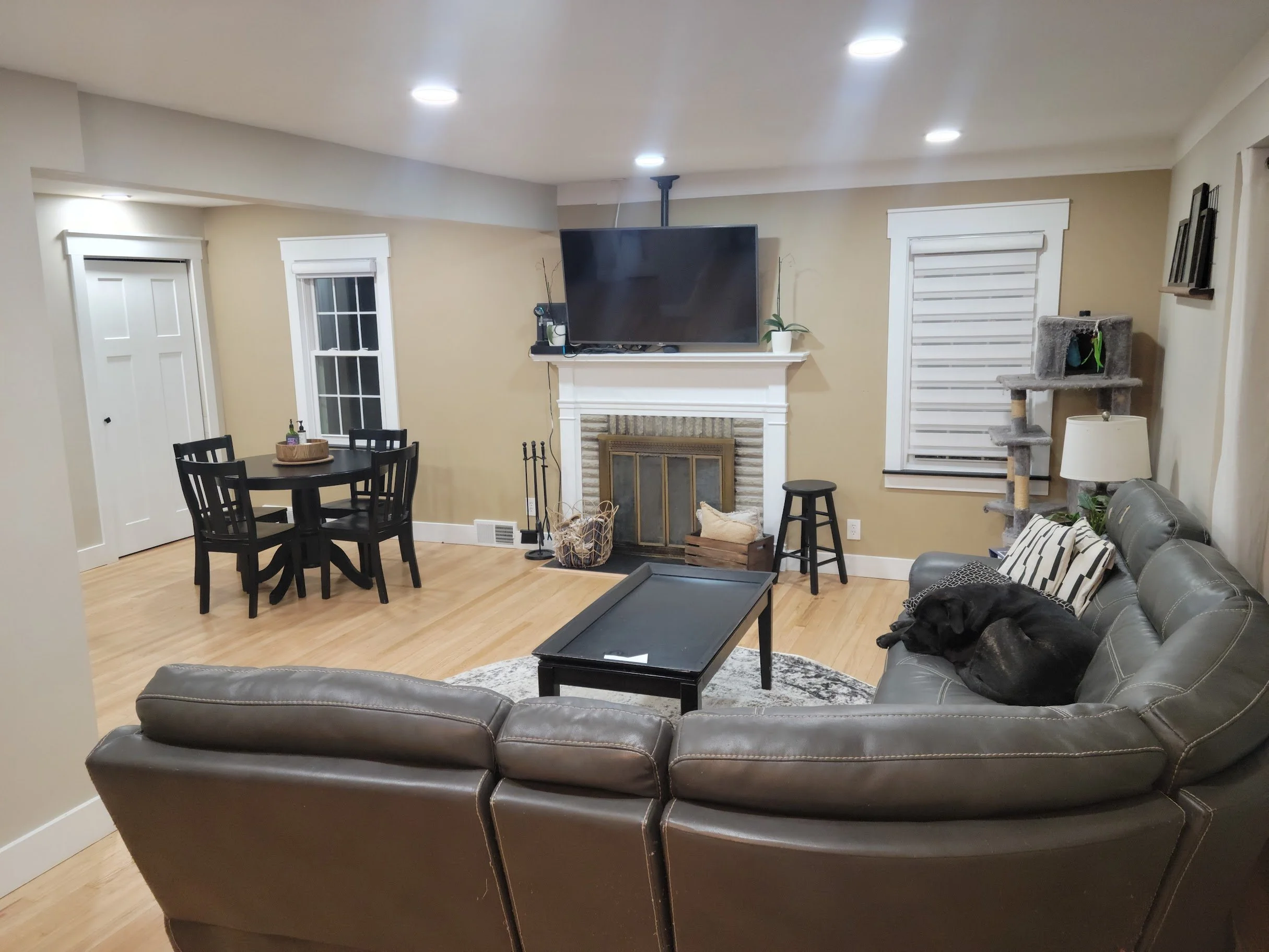 Living room with gray leather sofa, black dining table with chairs, fireplace, wall-mounted TV, beige walls, white trim, and cat tree near the window.