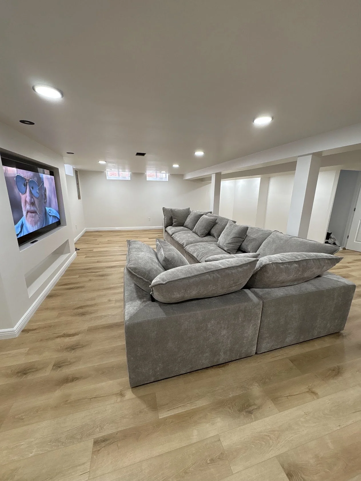 A basement living room with a large gray sectional sofa, hardwood floors, and a wall-mounted flat-screen TV showing a man with sunglasses.