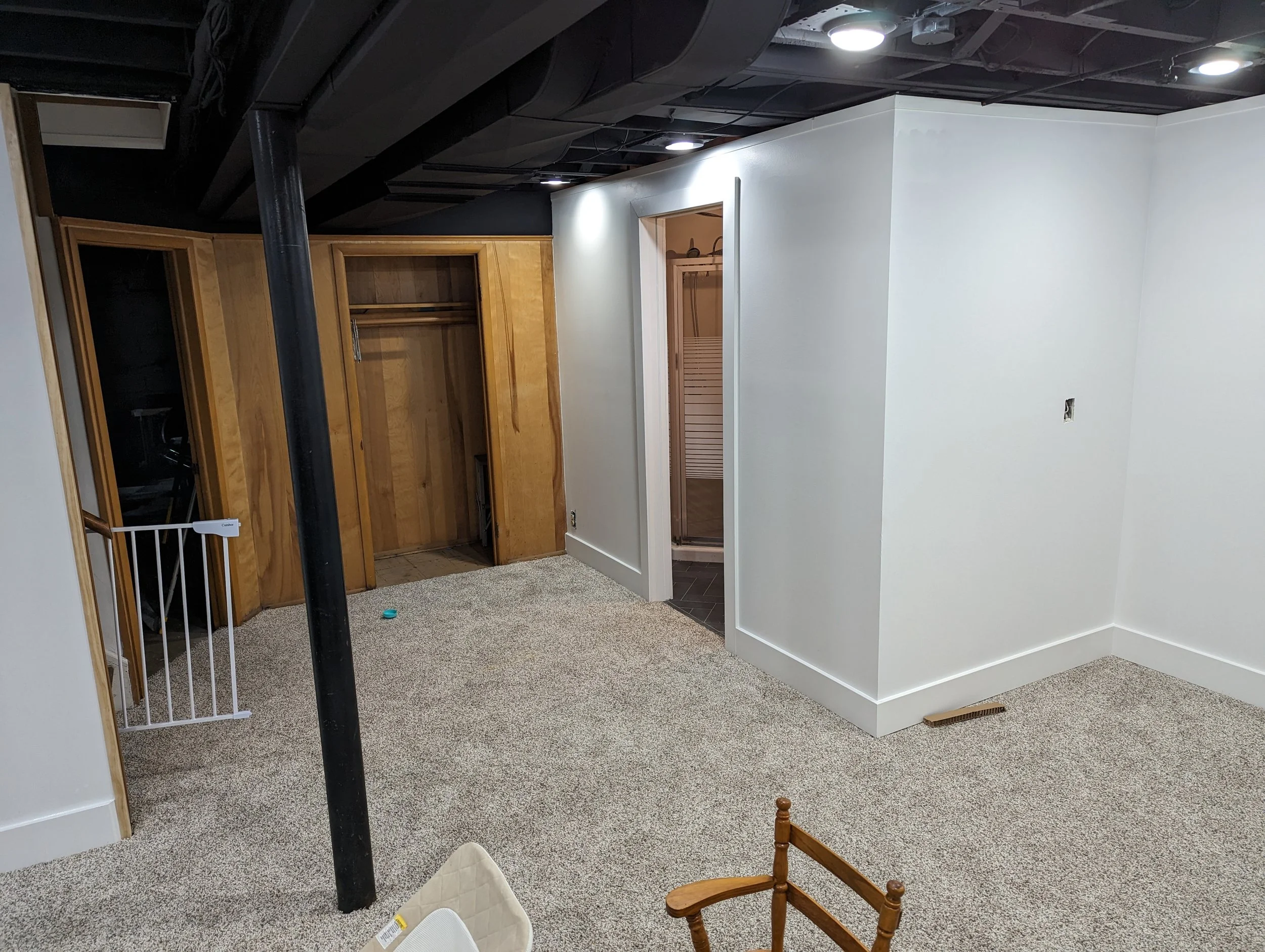Unfinished basement with white walls, black ceiling, and beige carpet. There is a wooden closet with an open door, a small bathroom with a doorframe, a black pole in the foreground, and some furniture including a small desk and a wooden chair.