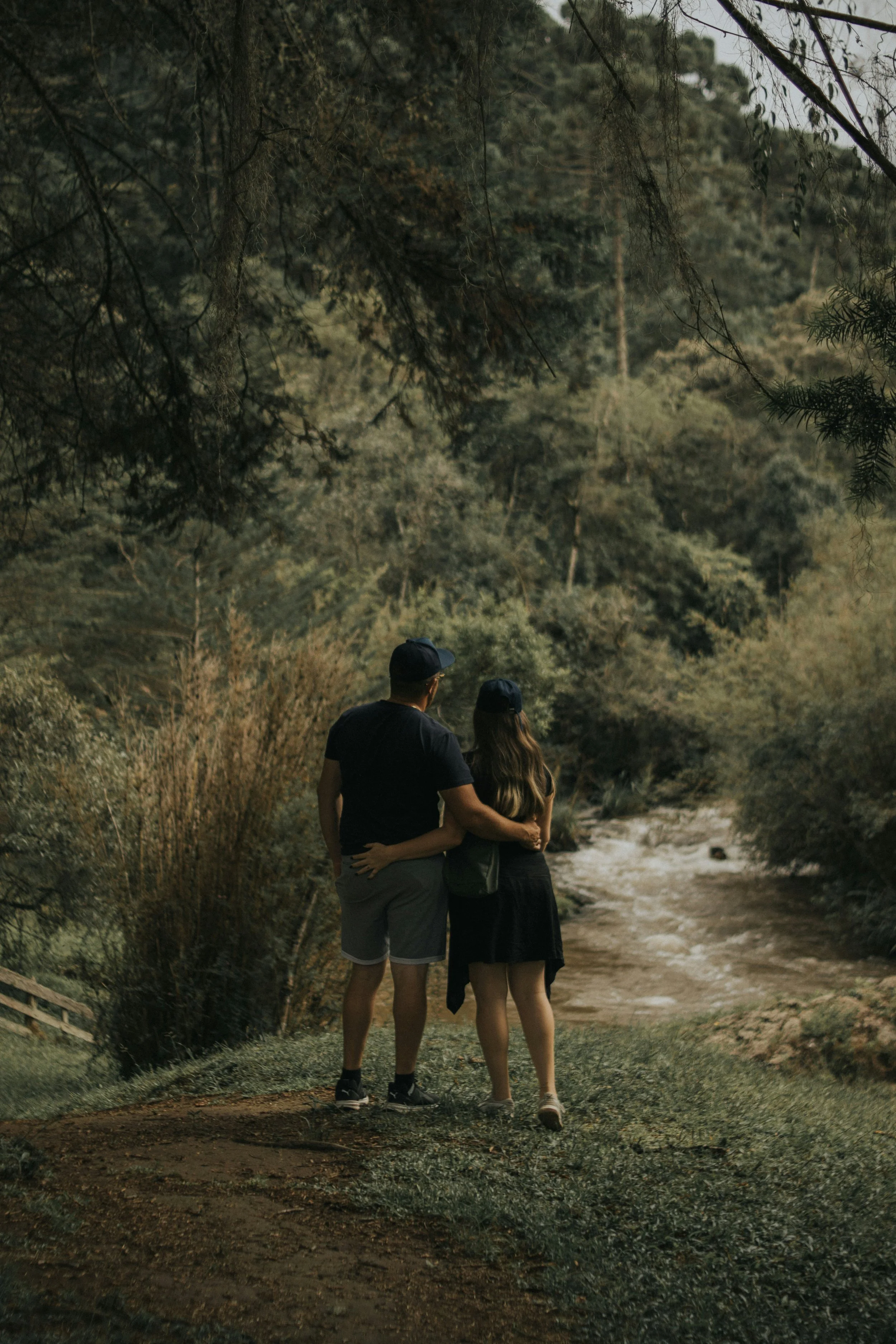 A man and a woman standing together on a nature trail surrounded by trees and bushes, with their arms around each other, under a canopy of leaves, during daytime.