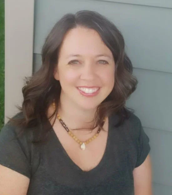 Smiling woman with dark wavy hair wearing a black top and a yellow beaded necklace, standing outdoors against a blue wall.