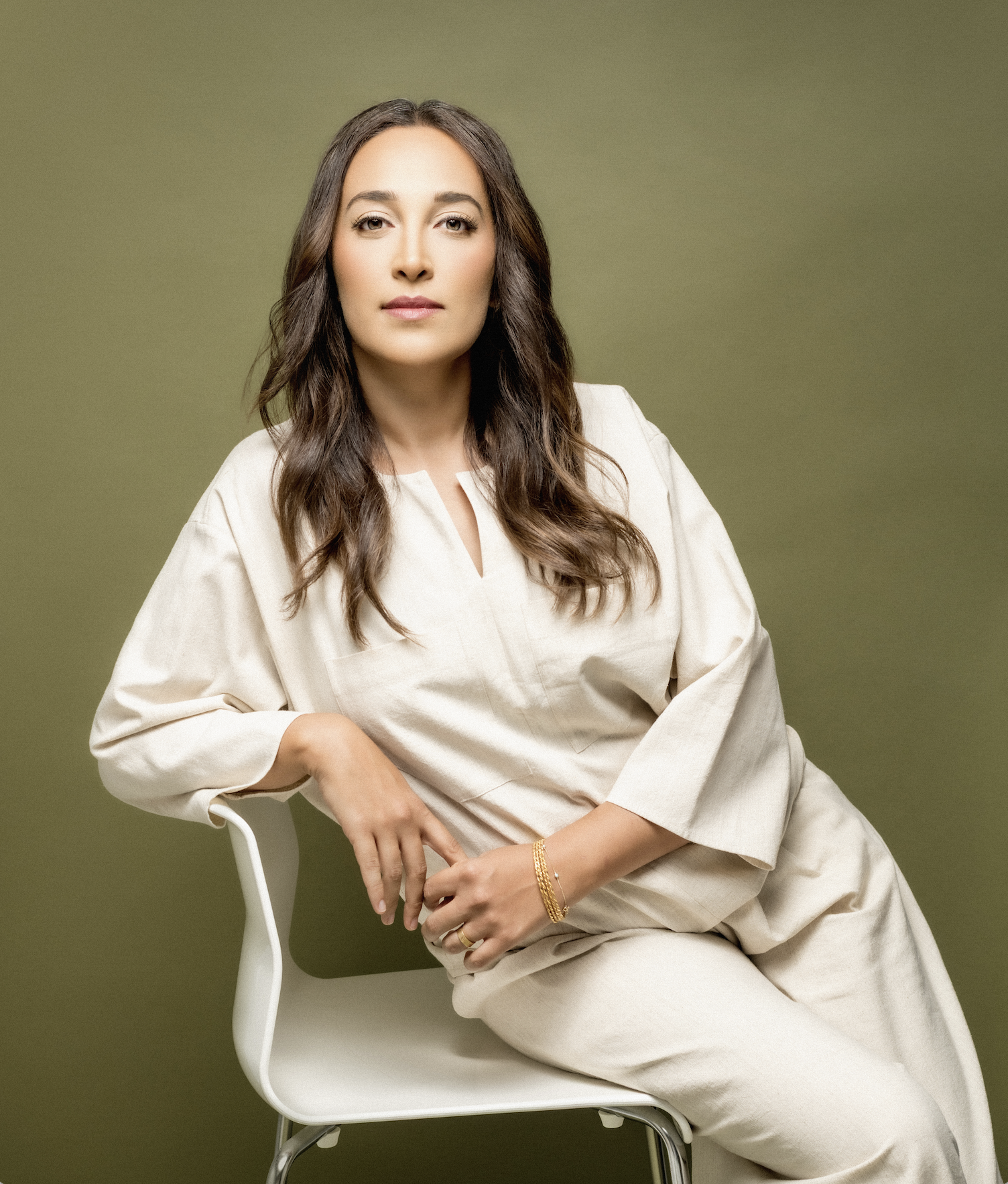 A woman with long brown hair, wearing a beige outfit, sitting on a white chair against a plain olive green background.