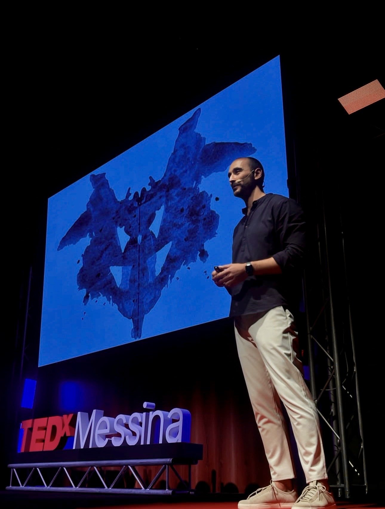 Dr. Mauro Cavarra presenting on stage at TEDx Messina with a large Rorschach inkblot test graphic displayed on the projector screen behind him.