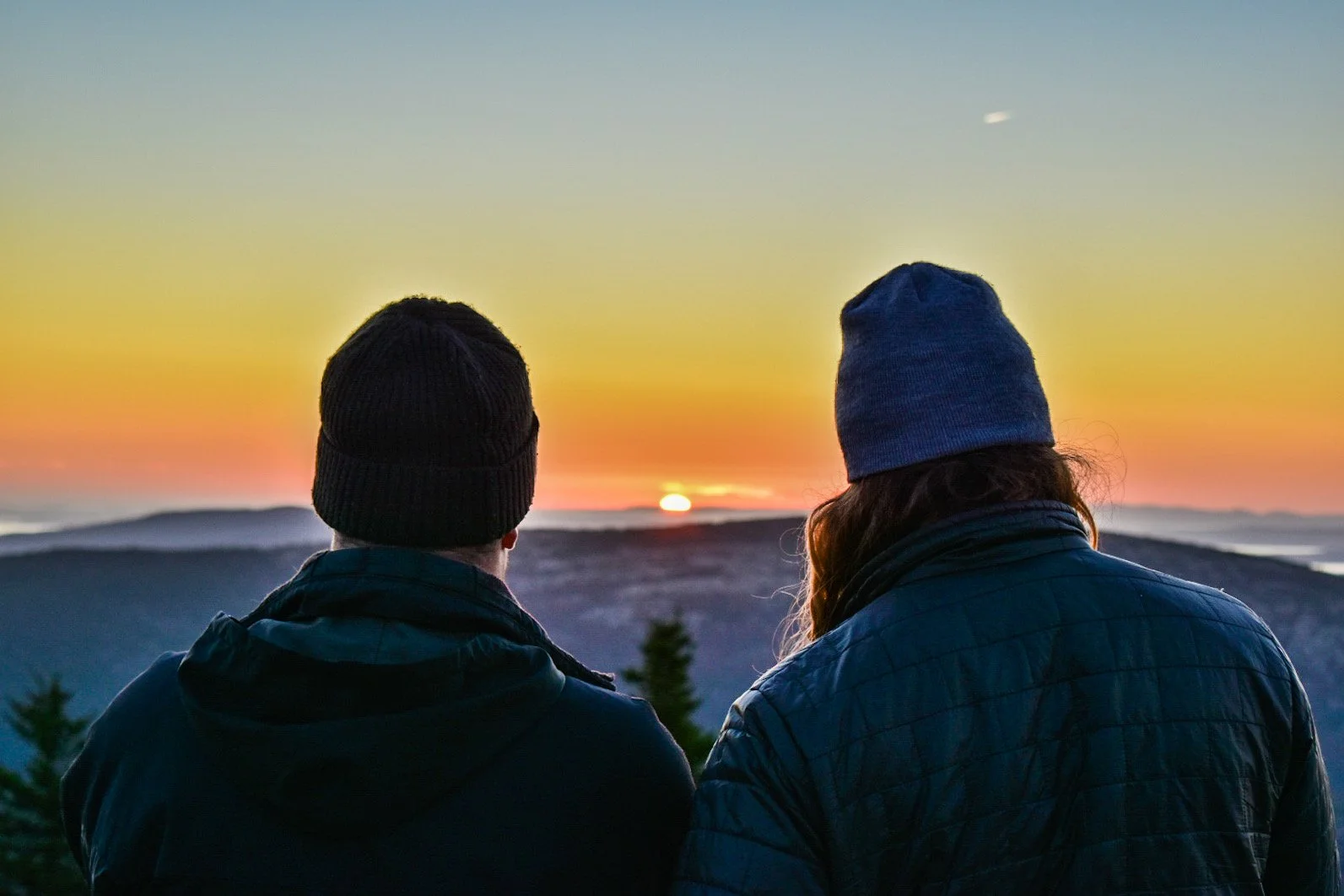 Two people wearing winter hats and jackets watching a sunset over mountains.