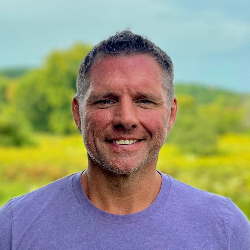 A smiling man with short dark hair and a beard, wearing a purple T-shirt, outdoors with green trees and cloudy sky in the background.