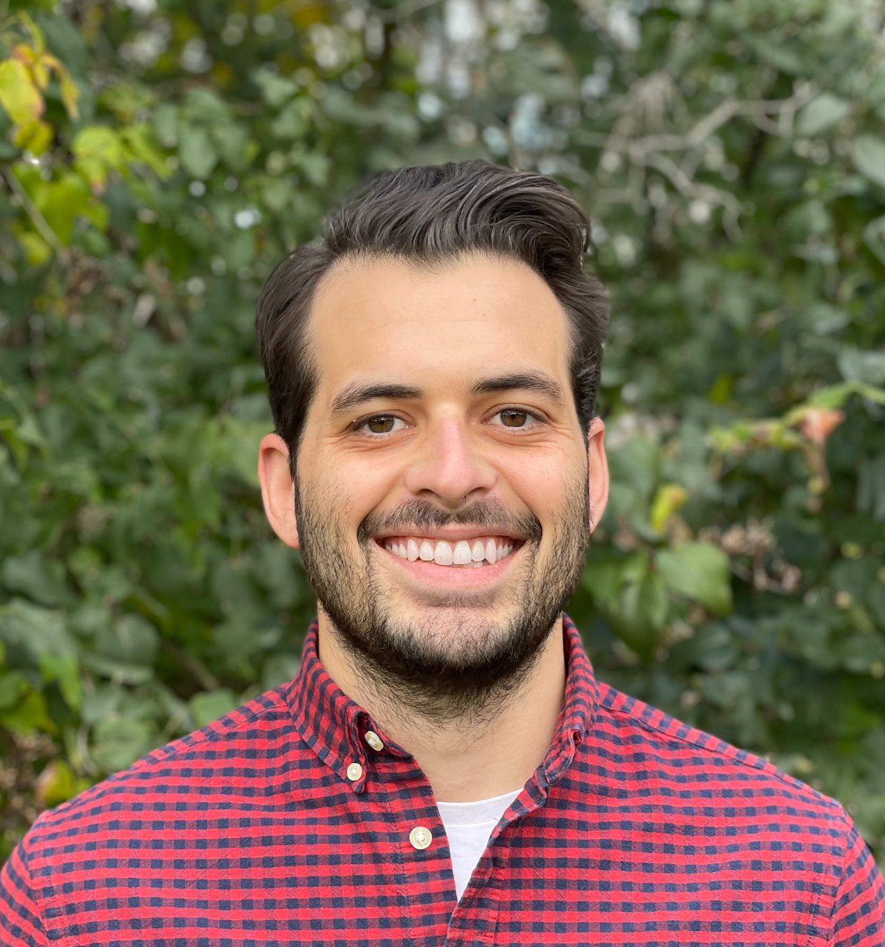 A smiling man with dark hair and a beard, wearing a red and black checkered shirt, standing outdoors in front of green foliage.