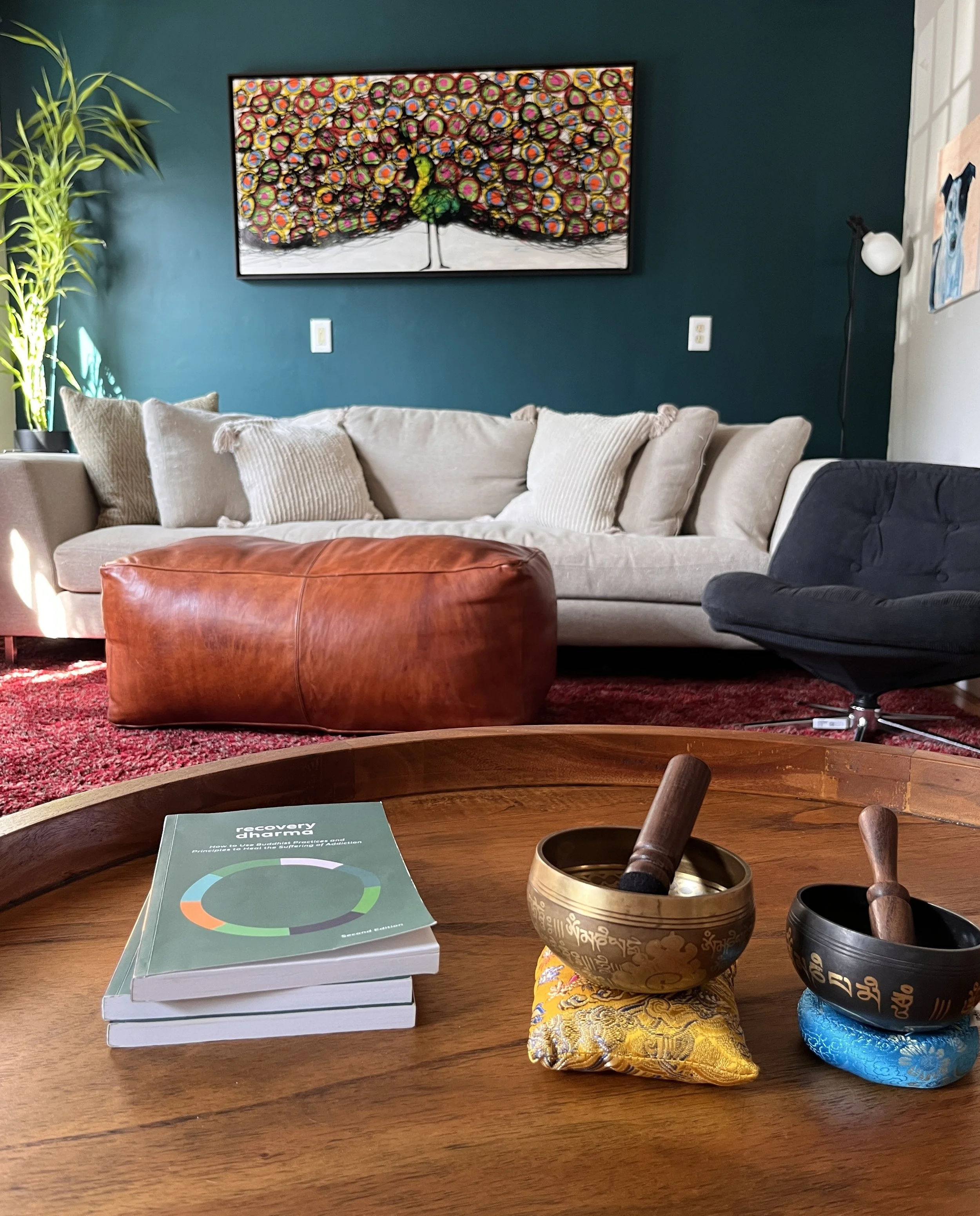 Interior of Common Collab's outpatient behavioral health office in Great Barrington, MA, featuring a comfortable living room with a beige sofa, leather ottoman, singing bowls, and a colorful abstract painting on a teal wall.