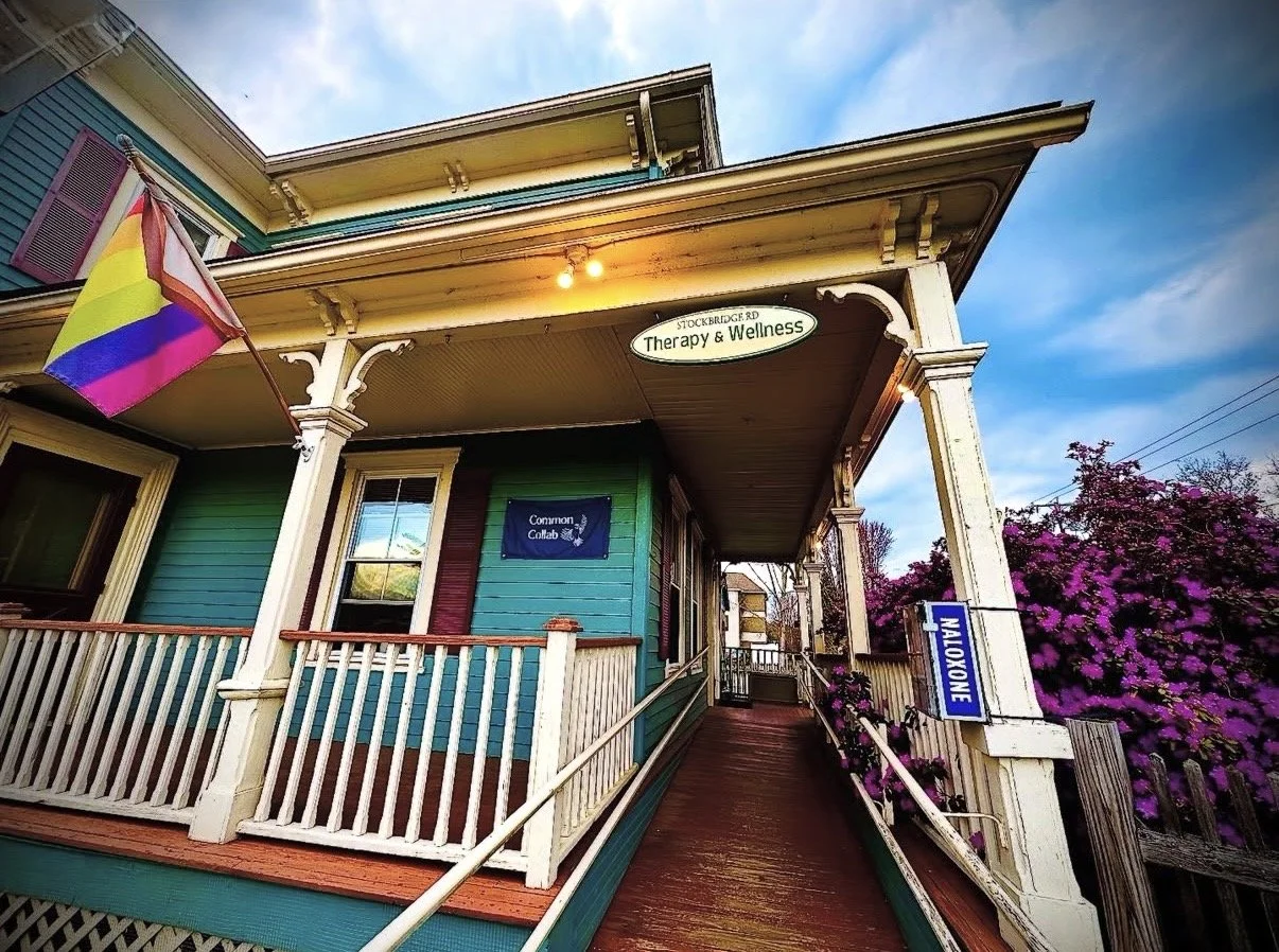Exterior of Common Collab at 405 Stockbridge Rd in Great Barrington, MA, a colorful Victorian building with a porch, rainbow pride flag, therapy and wellness signage, accessible ramp, and flowering plants.