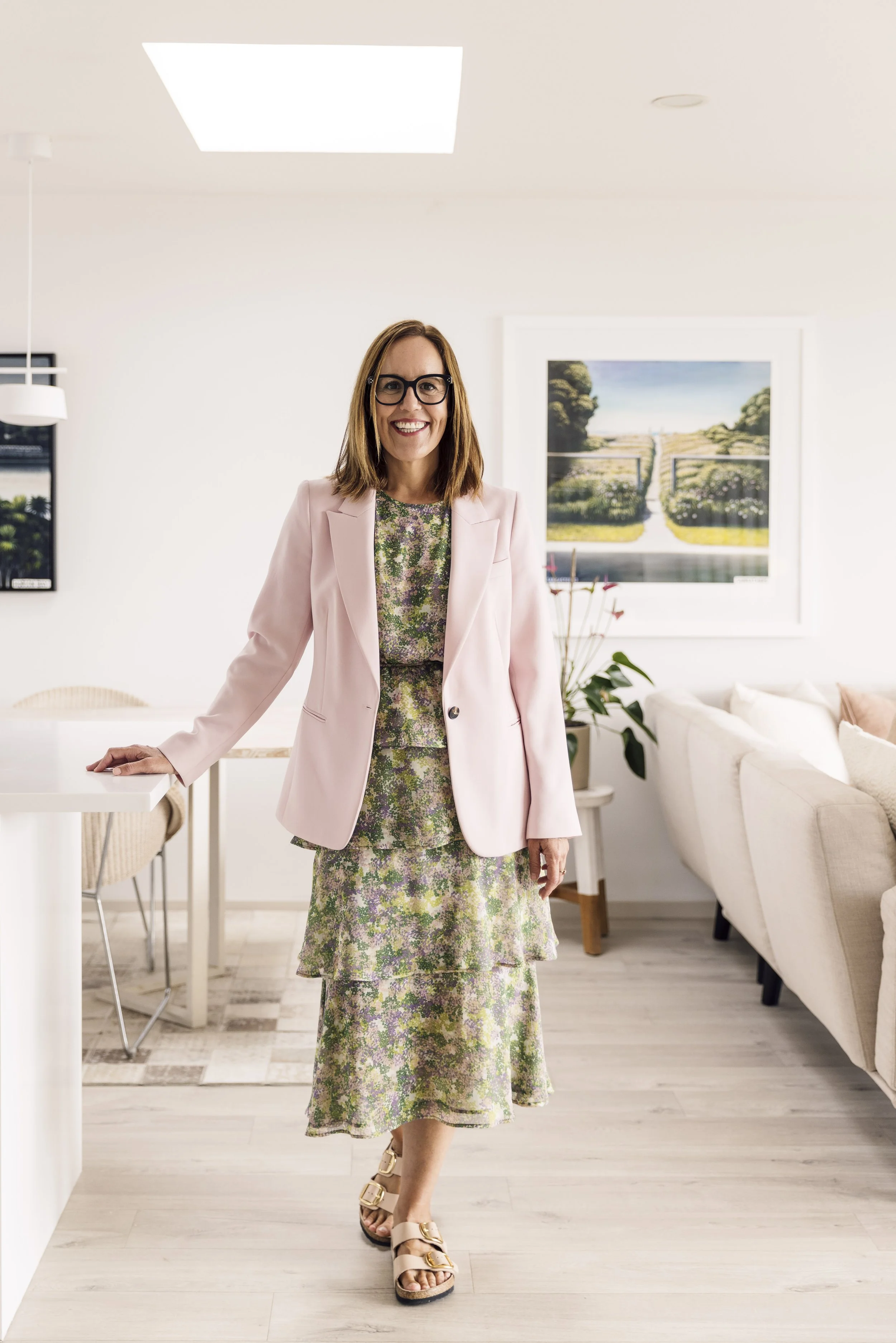 A woman with glasses smiling, wearing a pink blazer and a floral dress, standing in a bright, modern living room.