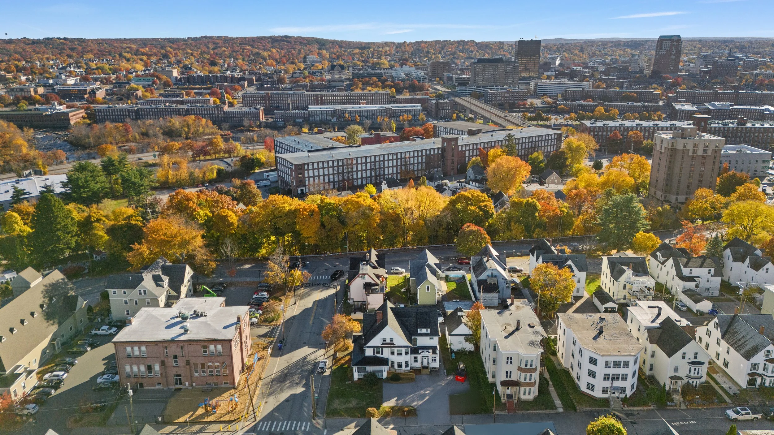 Aerial view of a city with colorful fall trees, residential houses, apartment buildings, and high-rise buildings under a clear blue sky.