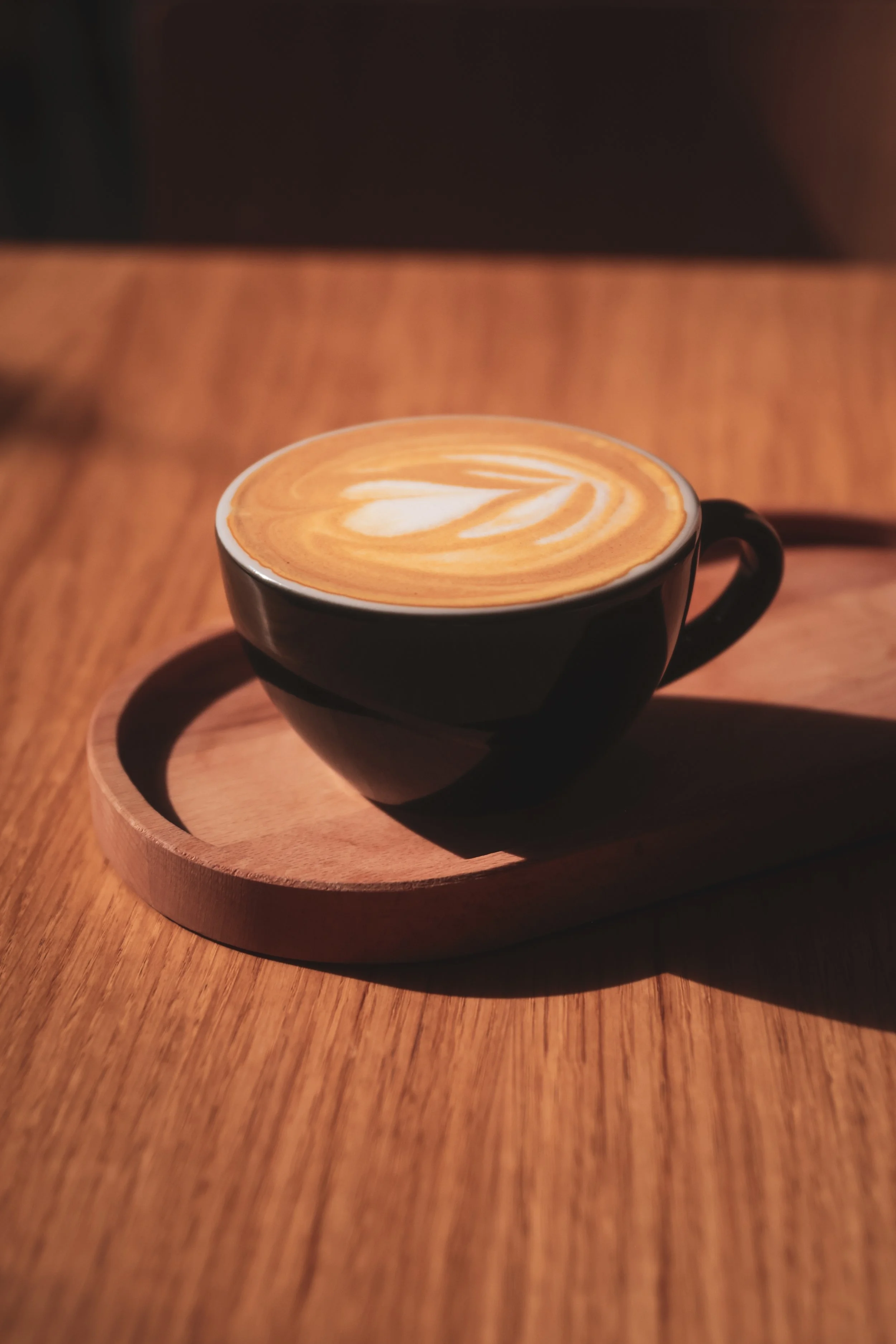 A black ceramic cup with a latte art heart design on top, sitting on a round wooden tray on a wooden table.