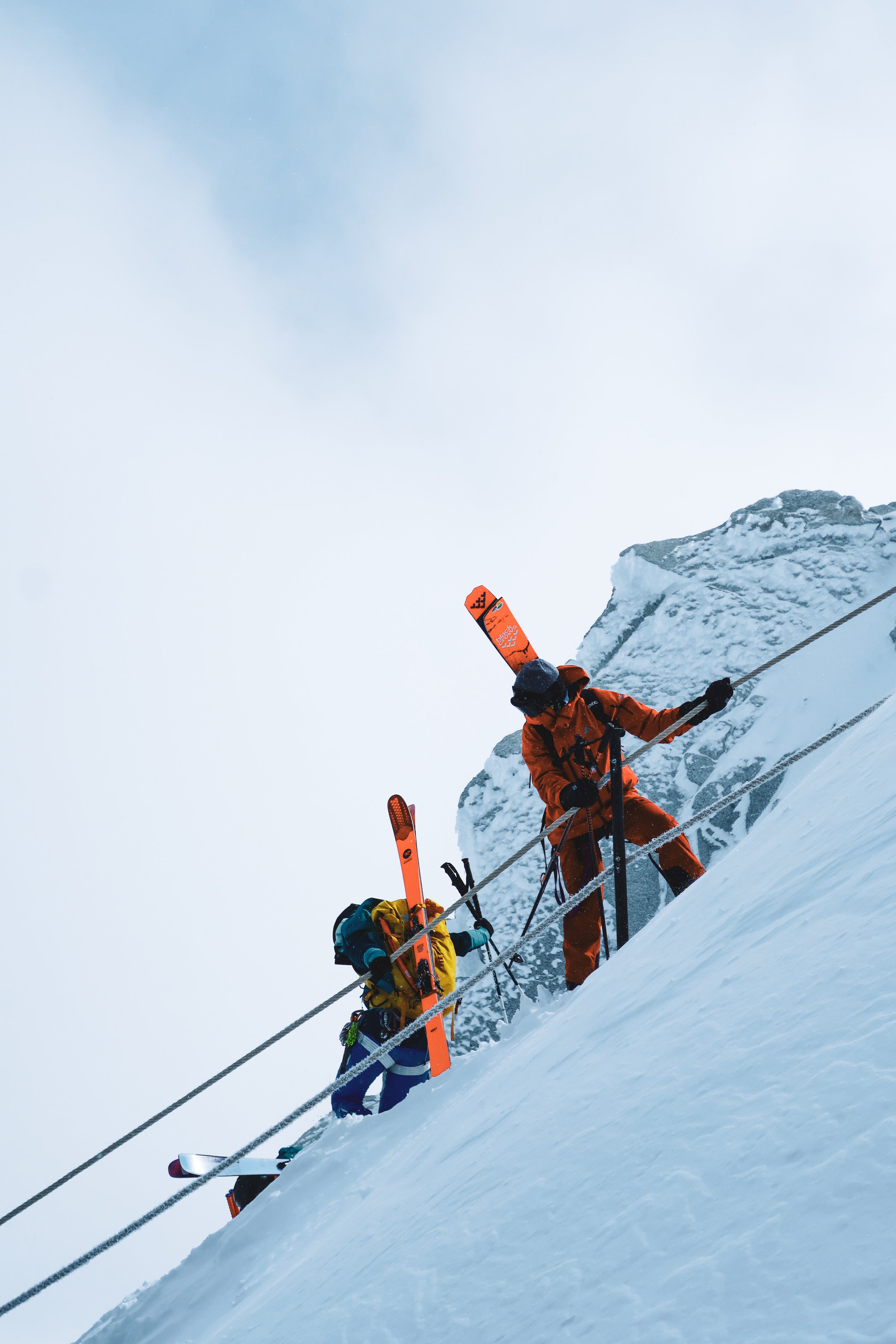 A group of mountaineers in bright orange and yellow jackets ascending a steep snow-covered slope with ice axes, wearing helmets and backpacks, against a cloudy, foggy mountainous background.