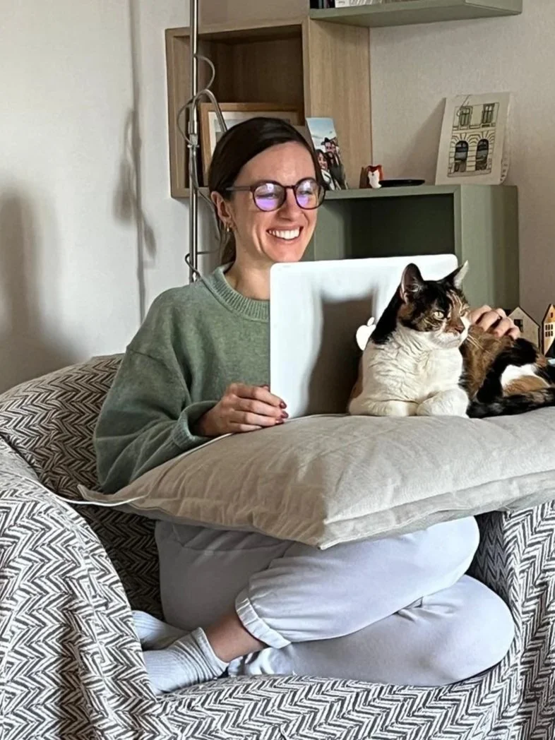 A woman with glasses and a gray sweater is sitting on a beige and black patterned couch, smiling while looking at a laptop. She is using a pillow to support a calico cat that is lying on her lap, looking to the right.