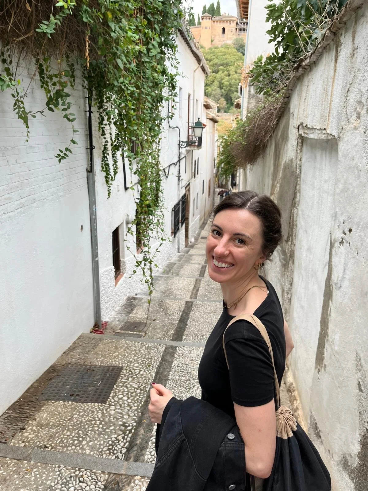 A woman with dark hair smiling in a narrow, stone-paved alleyway, with white walls on either side and greenery hanging from above. In the background, there is a historic building on a hill.