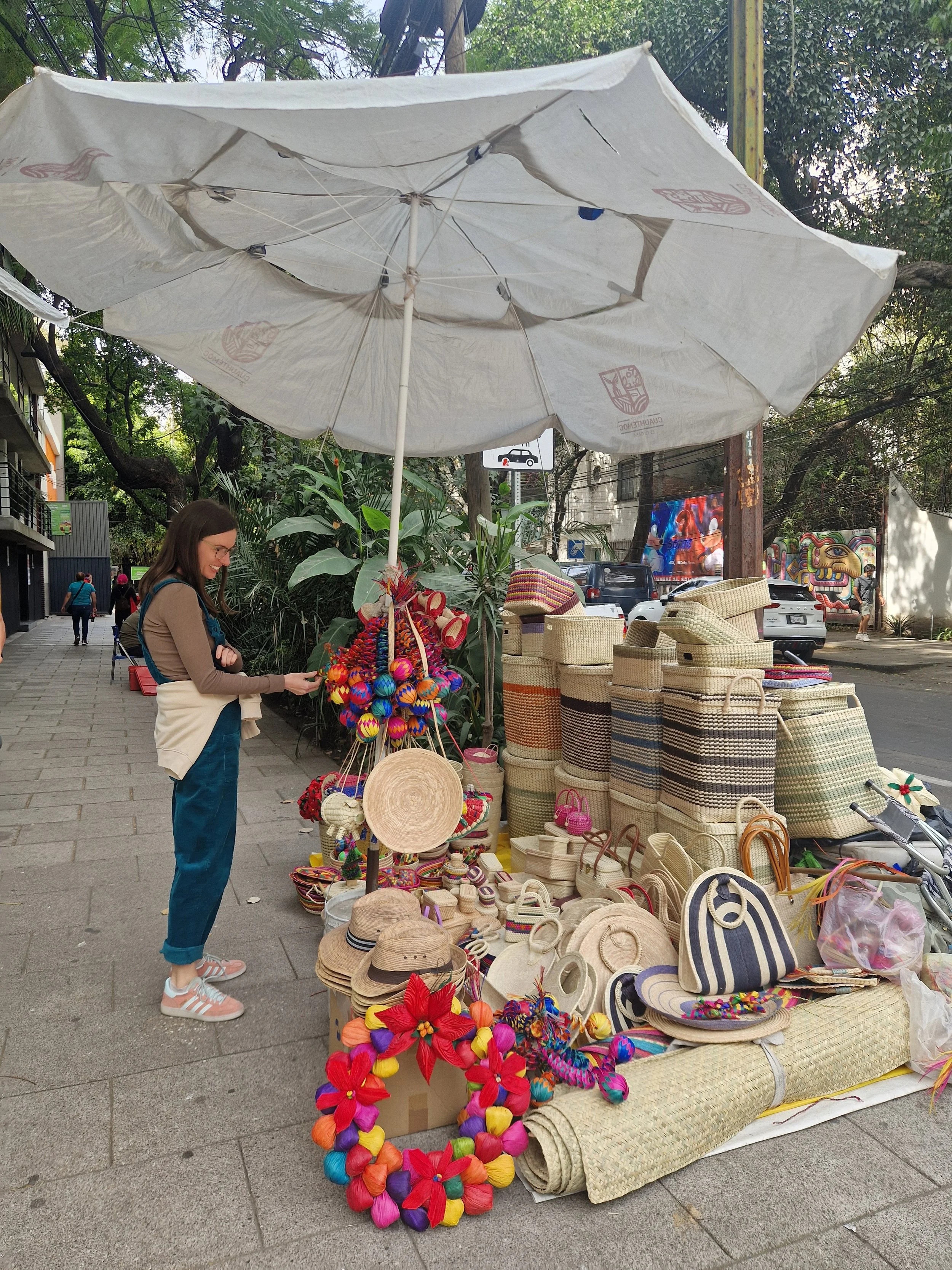 A street vendor selling colorful flower leis, straw hats, woven bags, and other accessories under a large white umbrella on a city sidewalk.