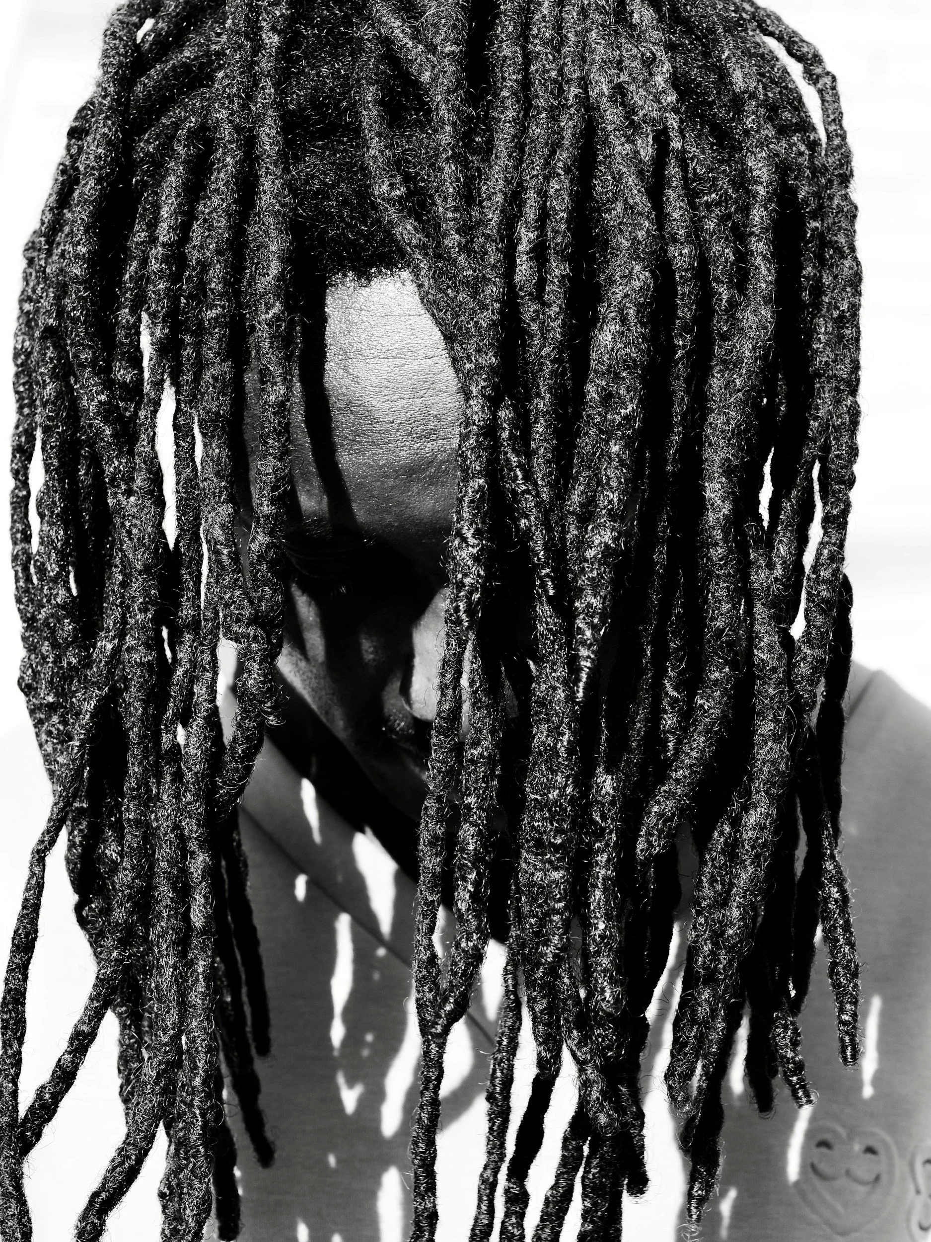 Close-up black-and-white photo of someone with long dreadlocks covering their face, with a shadowy background.