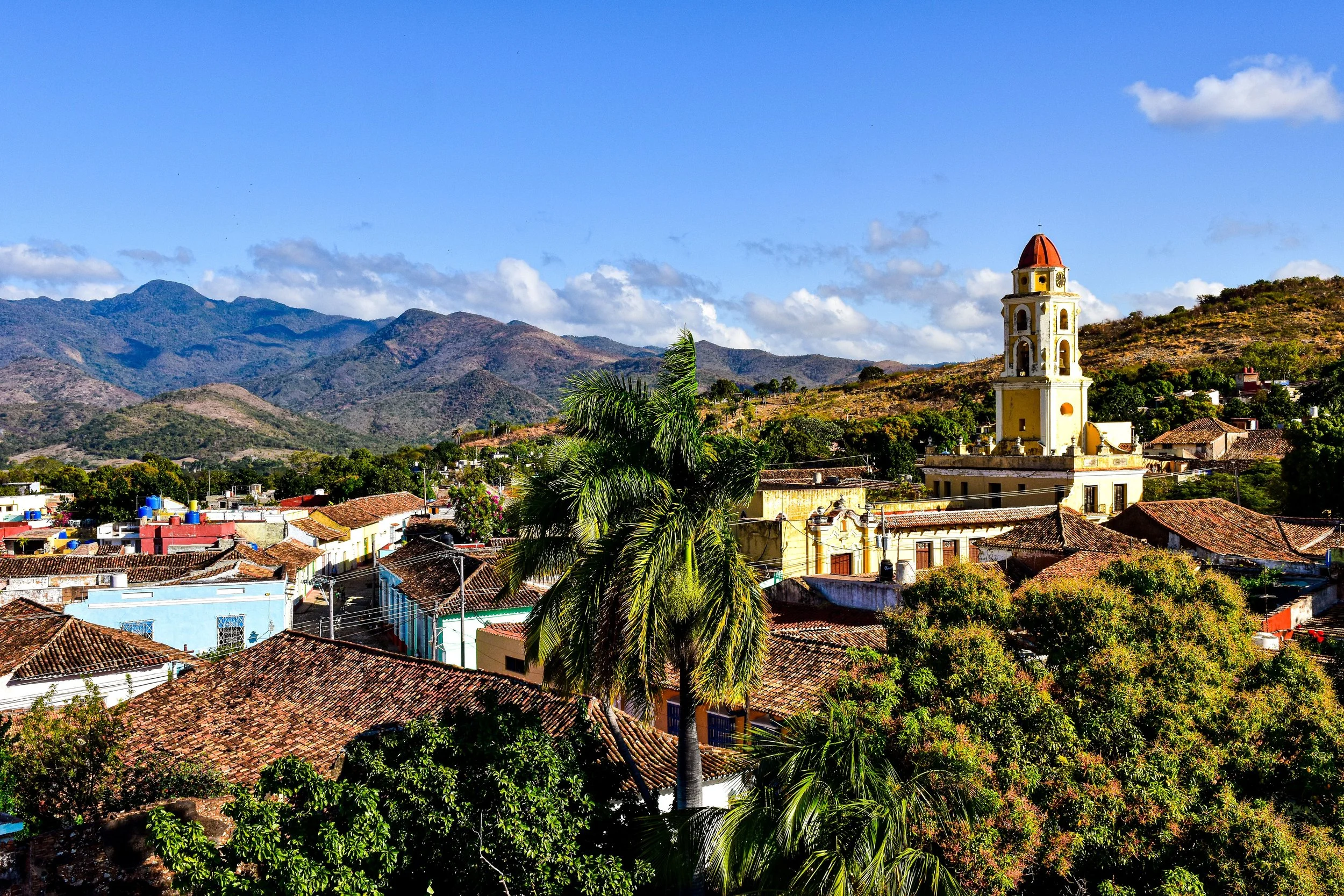 A picturesque town with red-tiled roofs, a prominent yellow church tower, lush green palm trees, and mountain ranges under a partly cloudy blue sky.