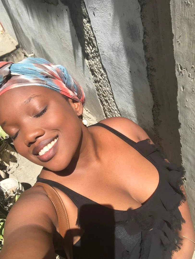 A woman with a colorful headscarf smiling with eyes closed, wearing a black top with ruffles, taking a selfie outdoors in sunlight.