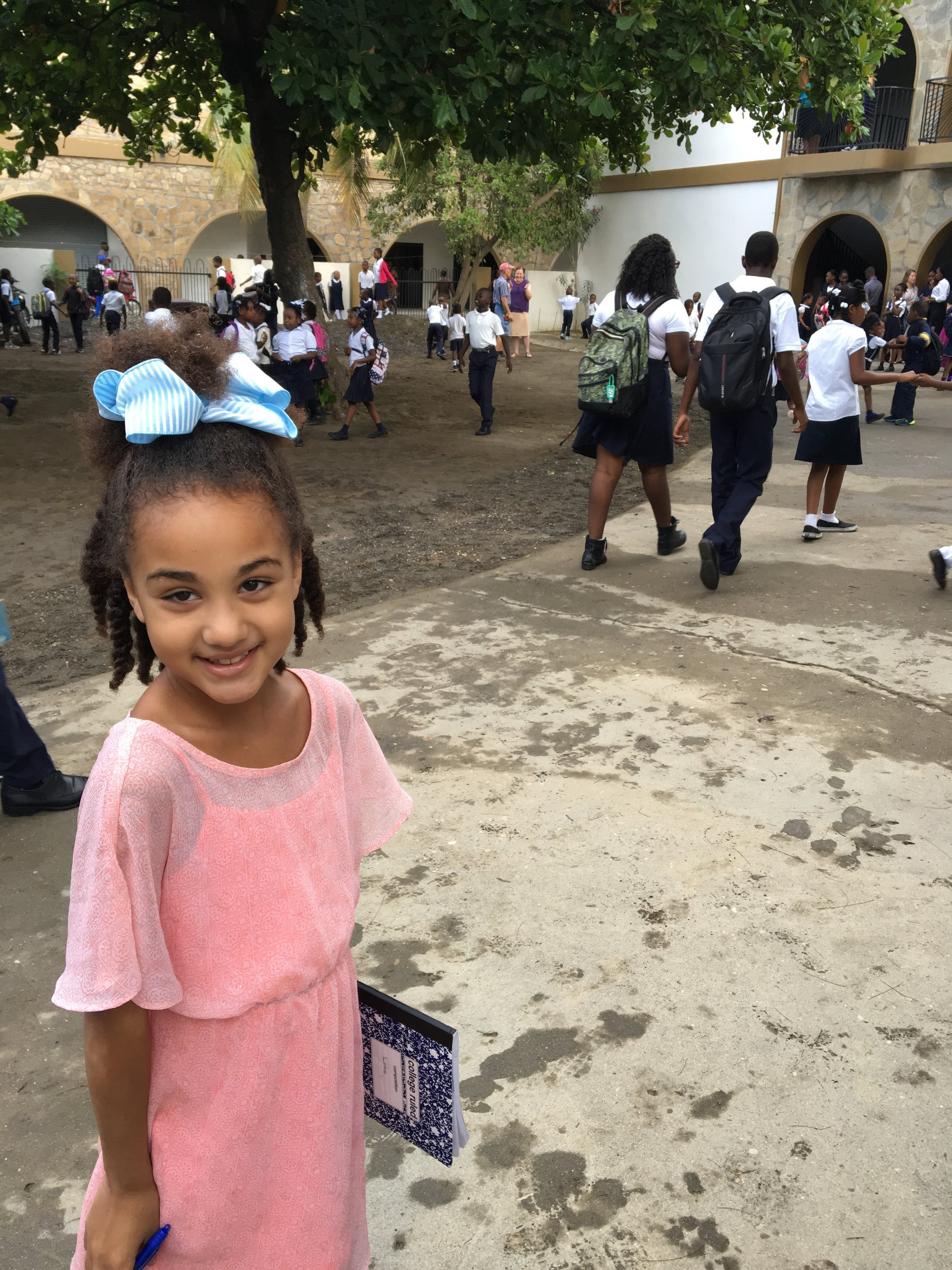 A smiling young girl with a large blue and white striped bow in her hair, holding a blue pen, standing outdoors in a schoolyard filled with students, teachers, and a large tree.