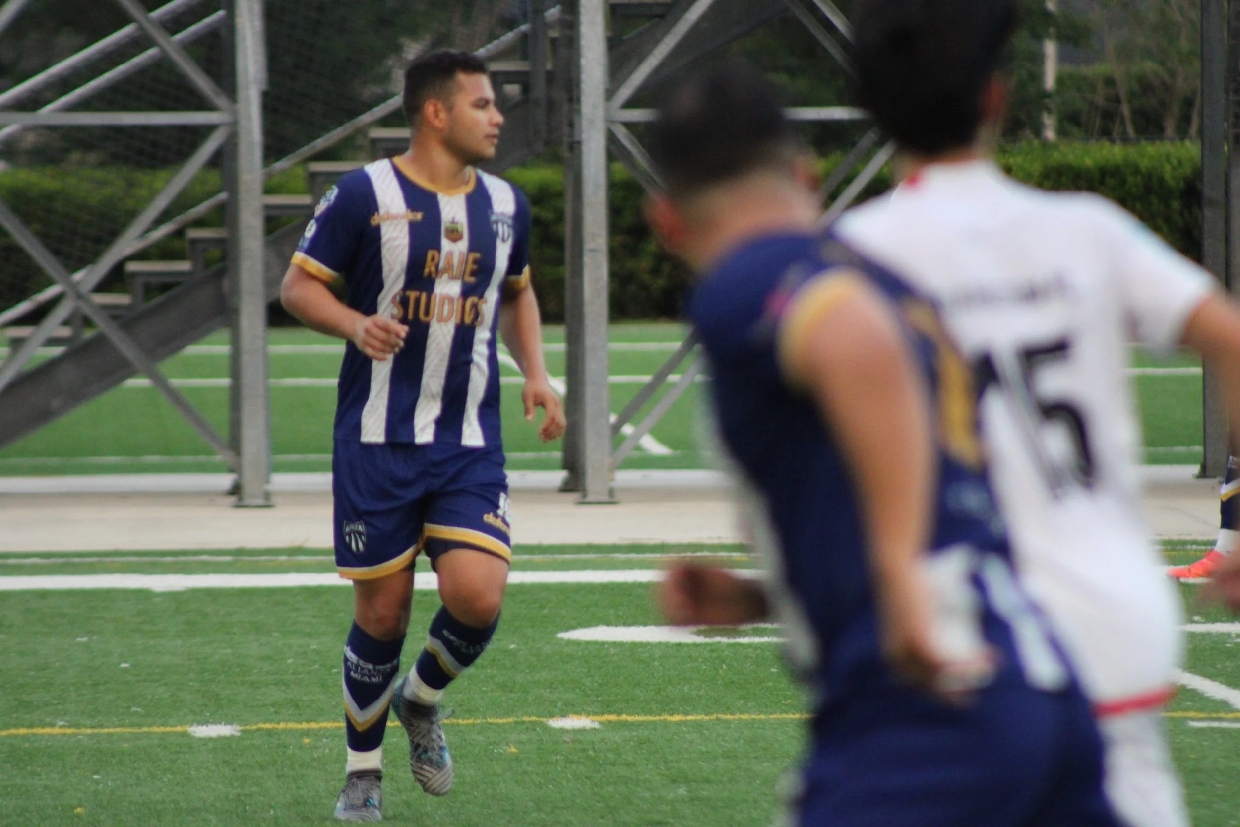 A young male soccer player in a blue and white striped jersey and shorts running on a green sports field with a metal staircase and greenery in the background, while other players are partially visible in the foreground.