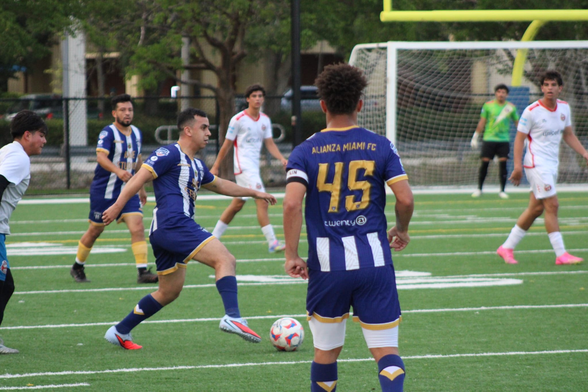 A group of young men playing soccer on a green football field during daylight. One player in a blue and yellow jersey labeled 'ALIANZA MIAMI FC' is in the foreground with his back to the camera, and others are visible in the background near the goal and fence.